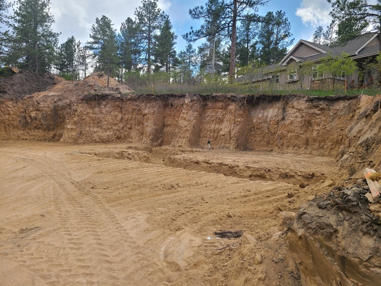 Excavated construction site with exposed soil and trees. A house is visible on the hilltop in the background.