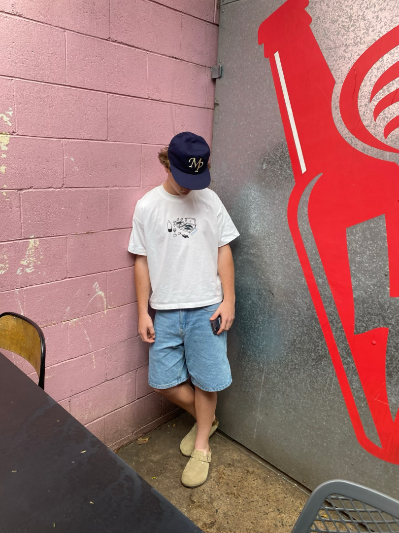 A young boy wearing a baseball cap, white T-shirt, and denim shorts is standing with his head bowed, holding a smartphone in one hand, next to a pink brick wall and a large red graphic on a gray wall.