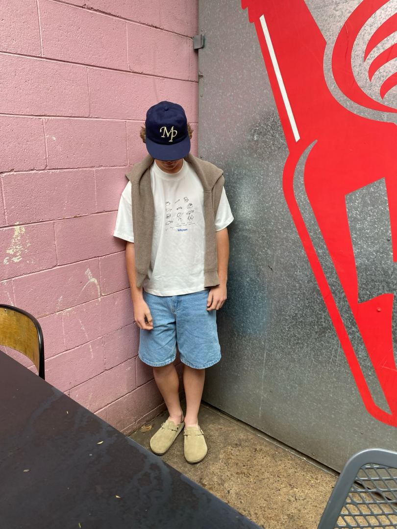 A young boy wearing a navy blue baseball cap, a white t-shirt, a beige hoodie draped over his shoulders, light blue shorts, and beige slip-on shoes standing in a corner with a pink brick wall on one side and a red and silver wall on the other side.