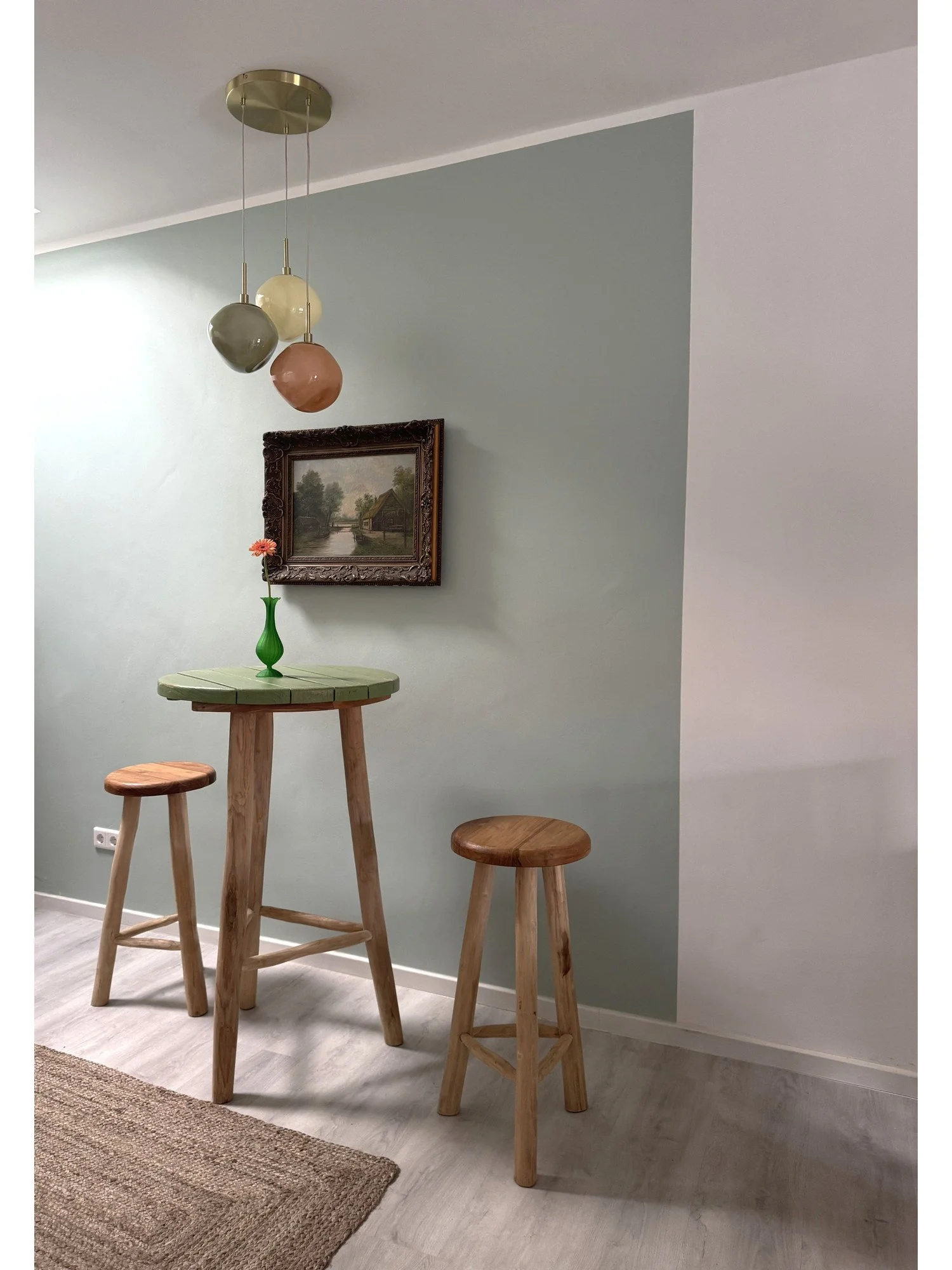 Ein Wohnzimmer mit einem kleinen Stehtisch und zwei Hockern aus Holz, eine grüne Vase mit einer roten Blume auf dem Tisch, ein gerahmtes Landschaftsbild an der Wand, moderne Deckenleuchte mit bunten kugelartigen Lampen, heller Holzboden und ein Teppich