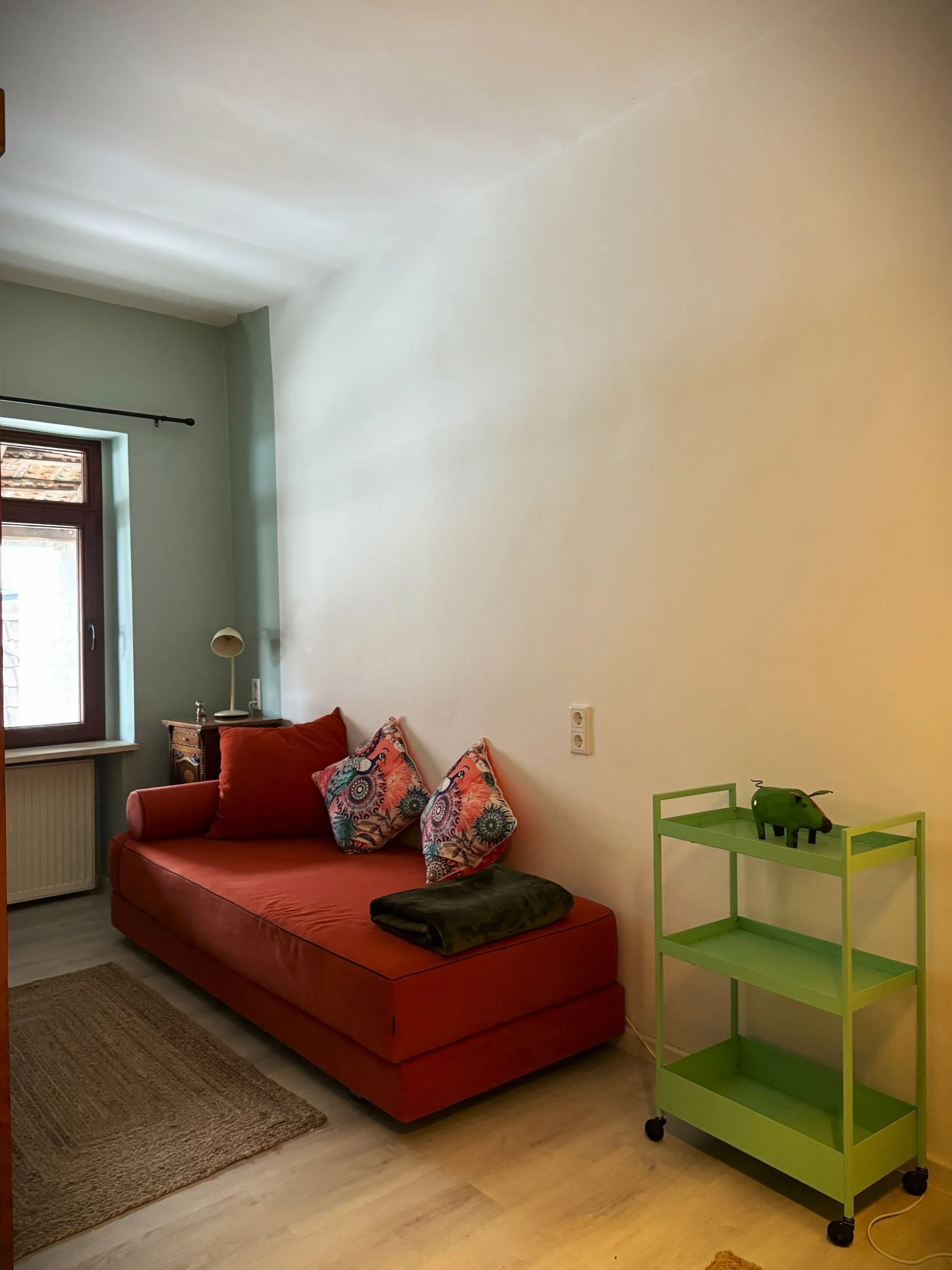 Ein Wohnzimmer mit einem roten Sofa, drei bunte Kissen, einem Fenster, einem kleinen Tisch mit Lampe, und einem grünen Rollwagen mit einer grünen Büffel-Figur auf der oberen Ablage.