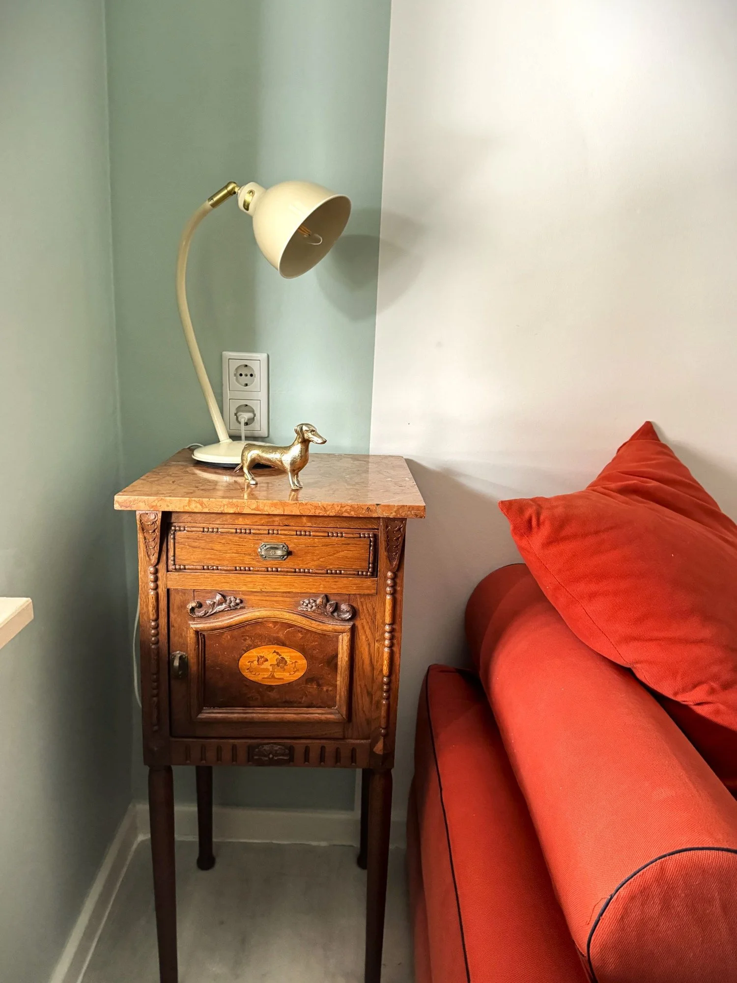 Ein Holznachtisch mit einer Marmortischplatte, einem kleinen goldfarbenen Dackelmodell und einer weißen Schreibtischlampe. Daneben ein rotes Sofa.