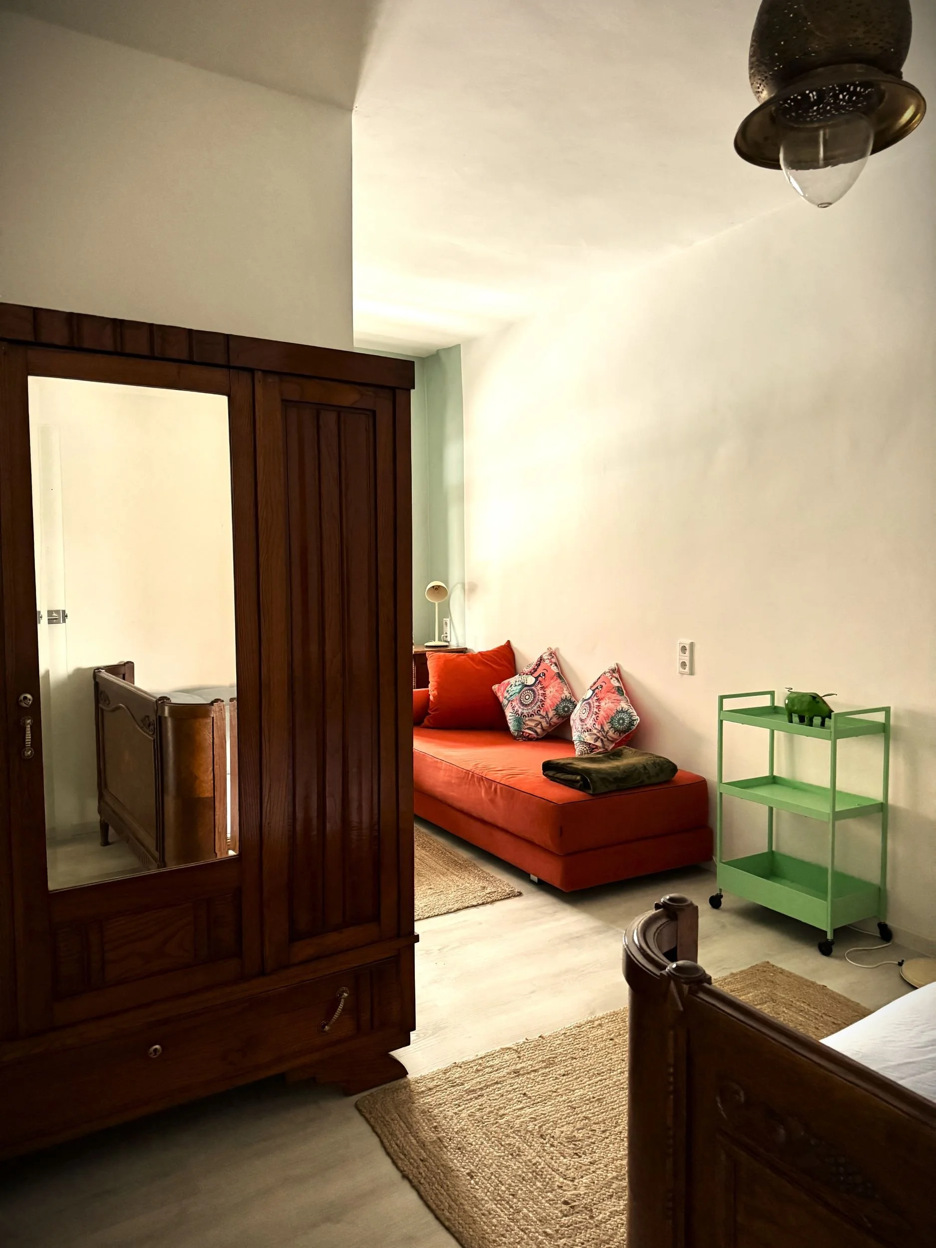 Ein Blick in ein Wohnzimmer mit einem roten Sofa mit bunten Kissen, einem grünen Beistelltisch mit einer Dekofigur, einem Holzmöbelstück vor weißer Wand und einem Holzschrank, warmer Beleuchtung.