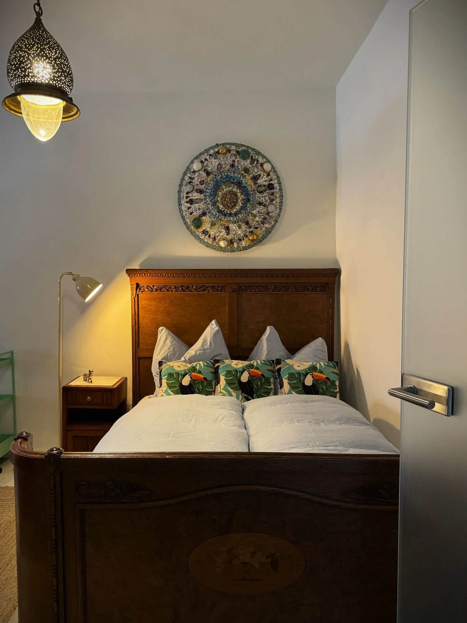 Ein Schlafzimmer mit einem Holzbett, drei Kissen mit Papageienmustern, einer Nachttischlampe, einem weißen Türgriff, einem Wandbild und einer Hängelampe.