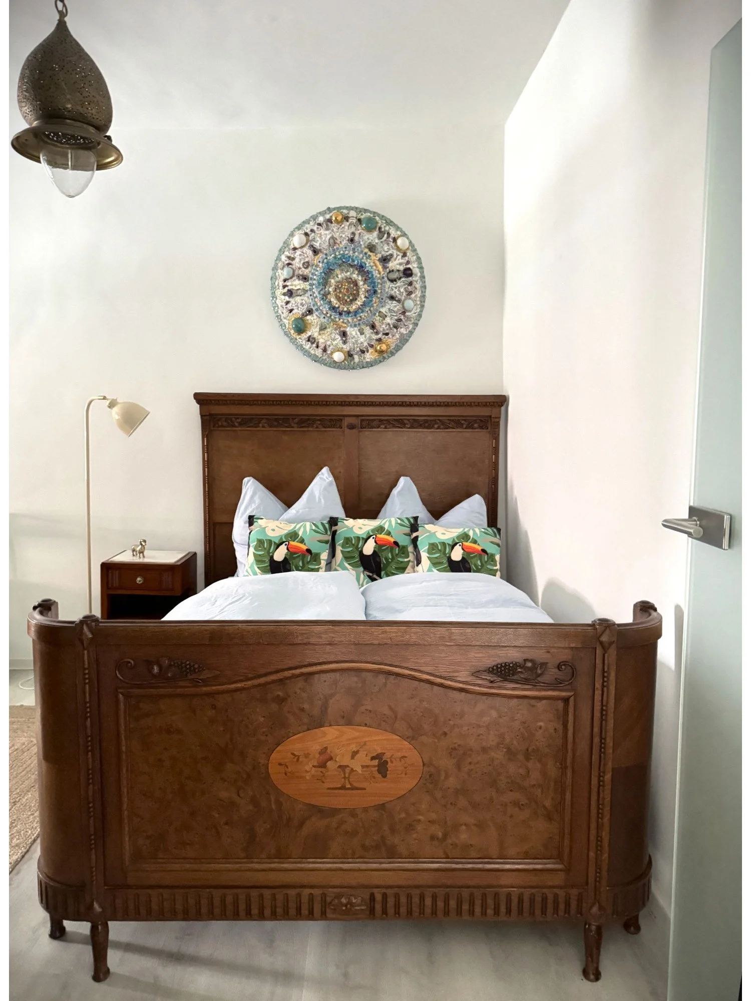 Ein Bett mit einem aufwändig geschnitzten Holzkopfteil, verziert mit einer kleinen bemalten Szene. Darauf liegen weiße Bettwäsche und Kopfkissen mit toucan-Motiven. Über dem Bett hängt eine Wanddekoration, die aus Muscheln und Perlen besteht. Rechts 
