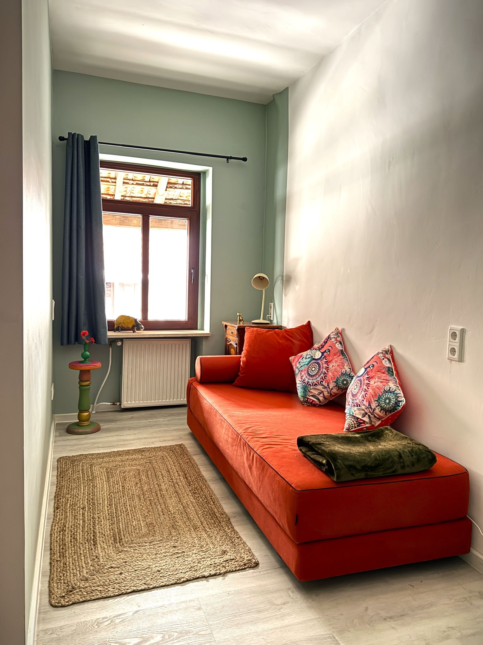 Ein kleines Wohnzimmer mit einem orangefarbenen Sofa, zwei bunte Kissen, ein brauner Teppich, ein Fenster mit dunkelblauen Vorhängen, ein kleiner Beistelltisch mit einer Vase und roten Blumen, ein Nachttisch mit einer Lampe, die Wandfarbe ist hellgrü