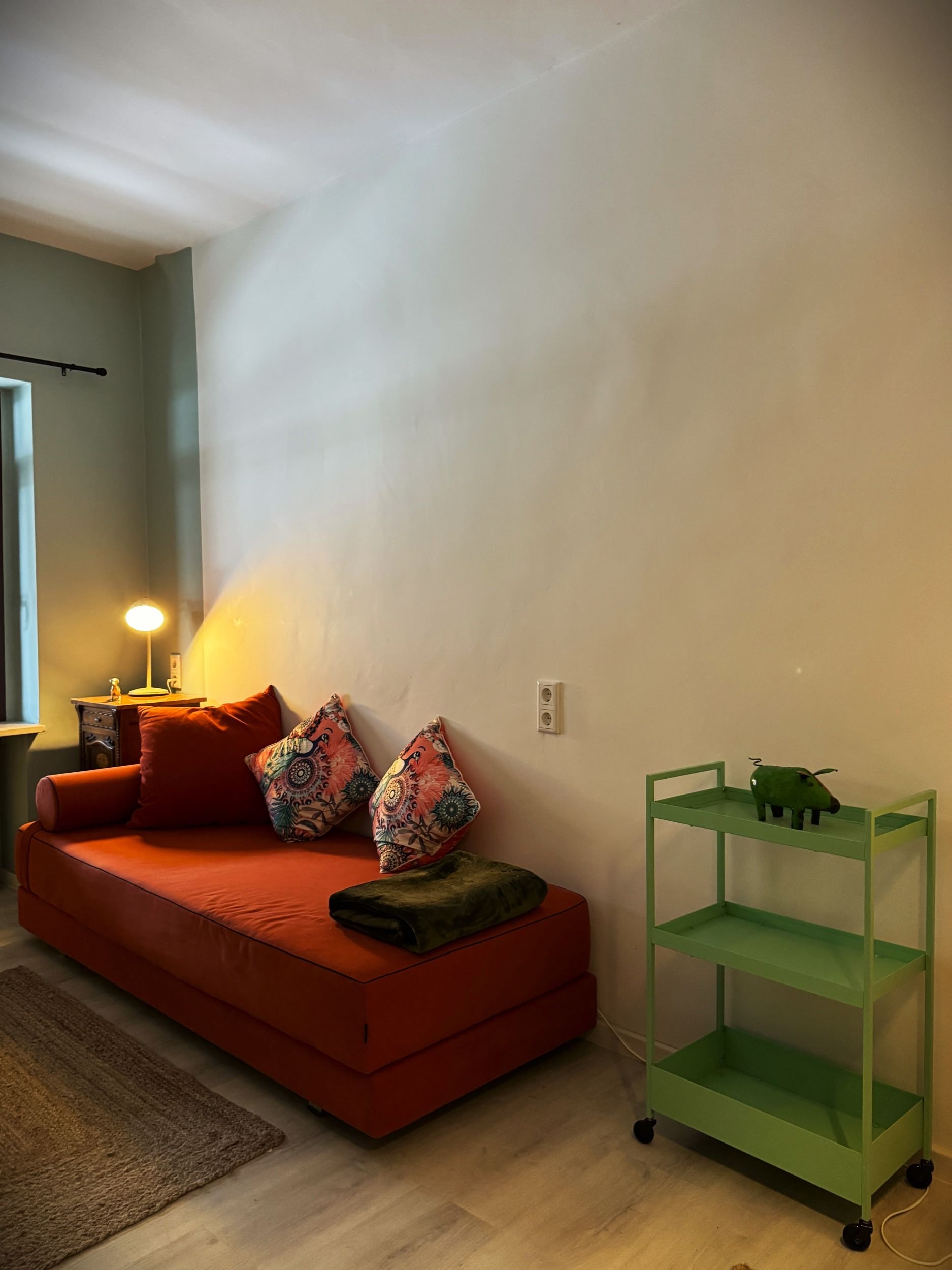 Ein Wohnzimmer mit rotem Sofa, bunten Kissen, einem kleinen Nachttisch mit Lampe, einem grünen Rollwagen mit einer grünen Elefantenfigur, einem Fenster mit schwarzen Vorhängen und einem Holzboden.