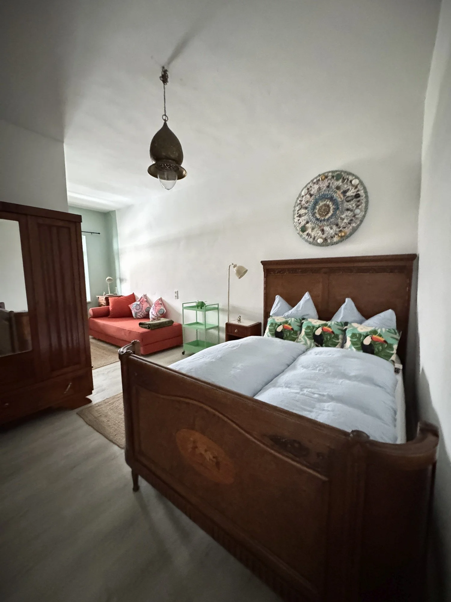  Schlafzimmer mit  Holz geschnitzten Bett, hellblauer Bettlaken und kissen mit Tukan-motiven, eine Nachttischlampe, einen grünen Wagentisch, eine Tagesdecke einem Einpersonenbett  im Hintergrund, sowie eine Kupferlampe.