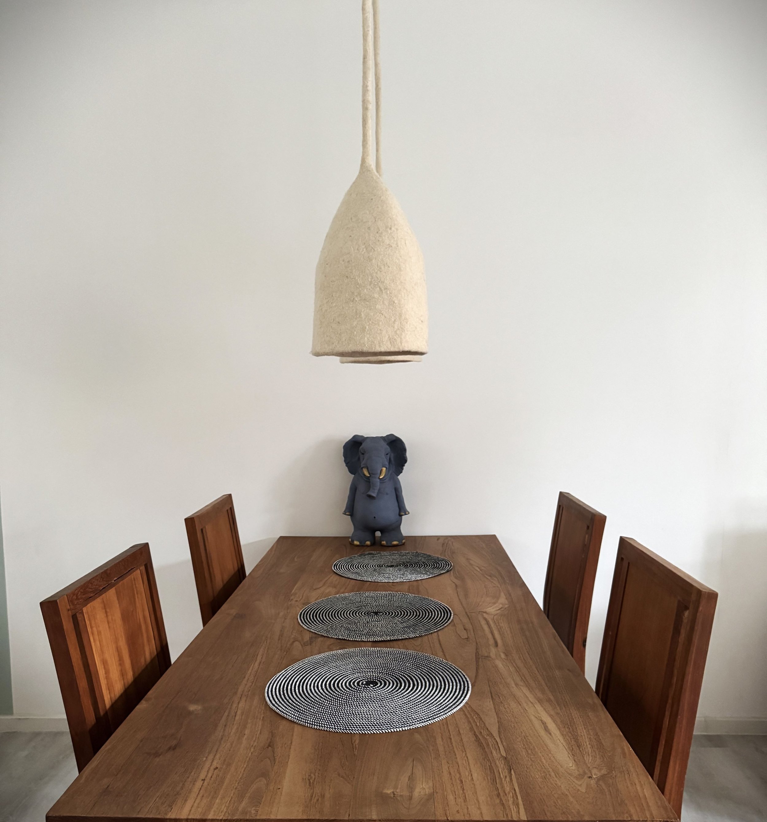 Ein Esstisch mit vier Holzstühlen, drei schwarzen Thunfisch-Teelichter, einer Elefantenfigur in der Mitte und einer großen, weißen Deckenlampe.
