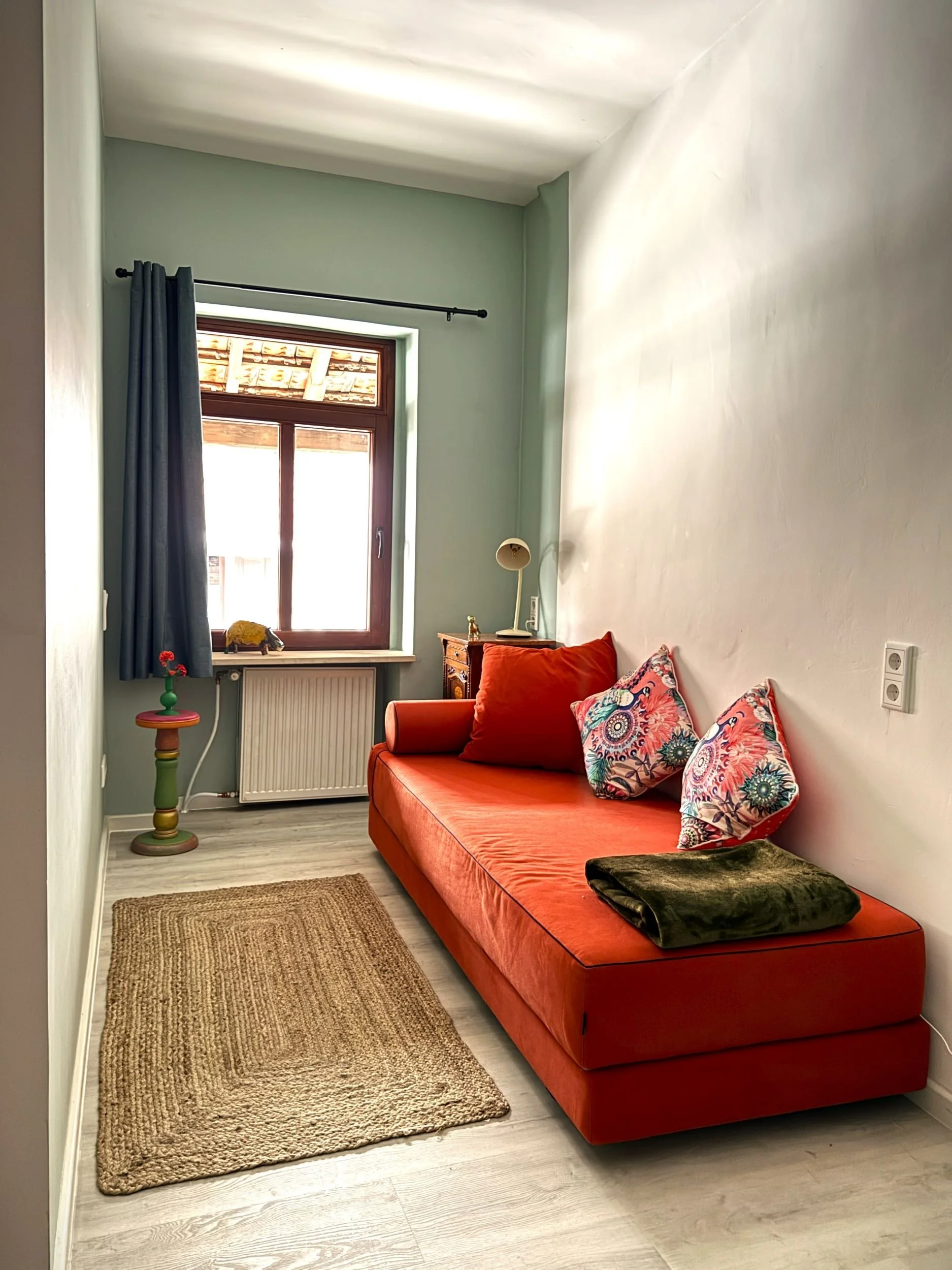 Ein kleines Wohnzimmer mit einem orangefarbenen Sofa, bunten Kissen, einem Fenster mit blauen Vorhängen, einem kleinen Teppich und einem kleinen Beistelltisch.