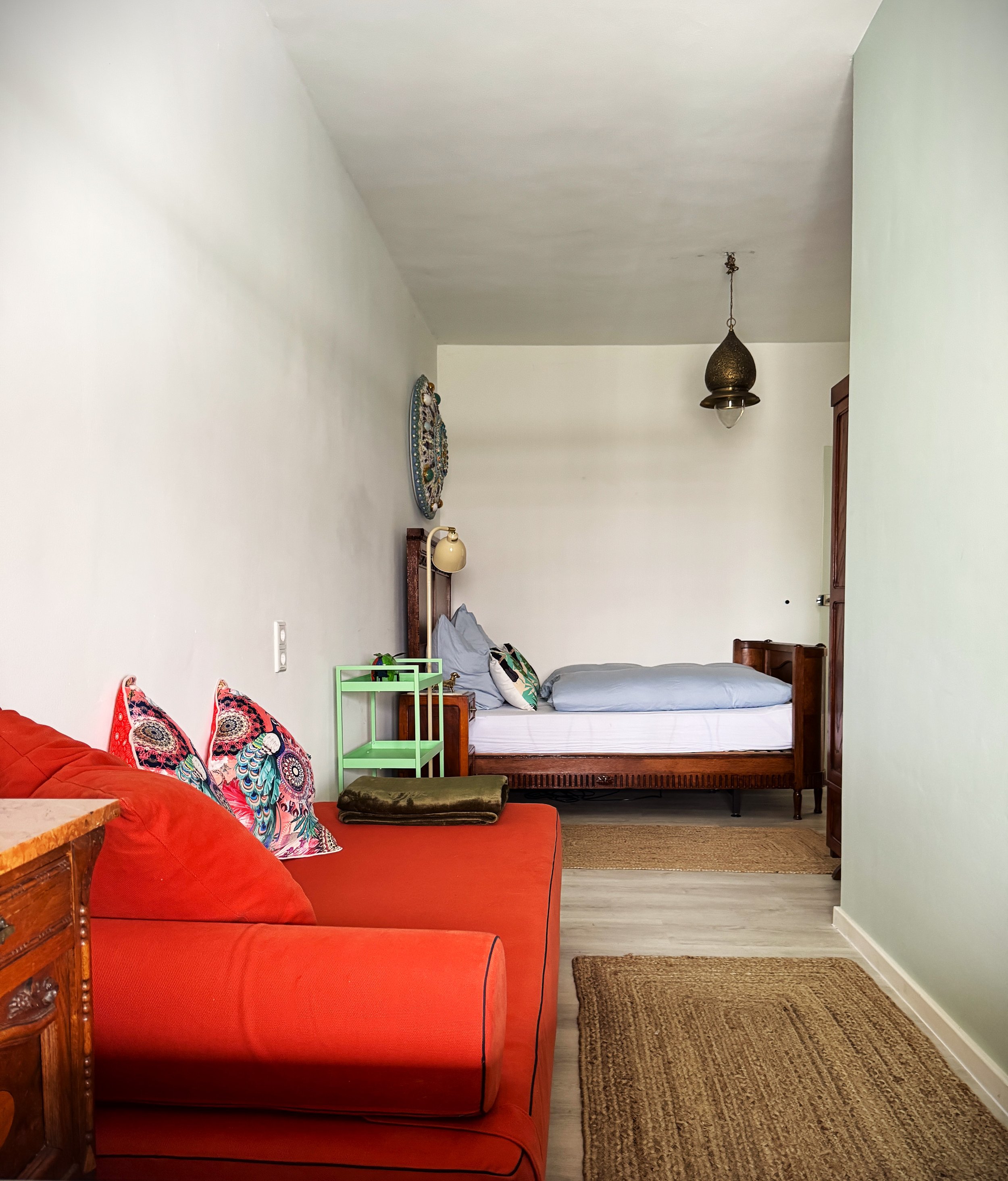 Schlafzimmer mit einem orangefarbenem Bett im dänischen Design, farbigen  Kissen,  Wagentisch, einem Einzelbett im Hintergrund, einer Kupferund Jugendstillbett.