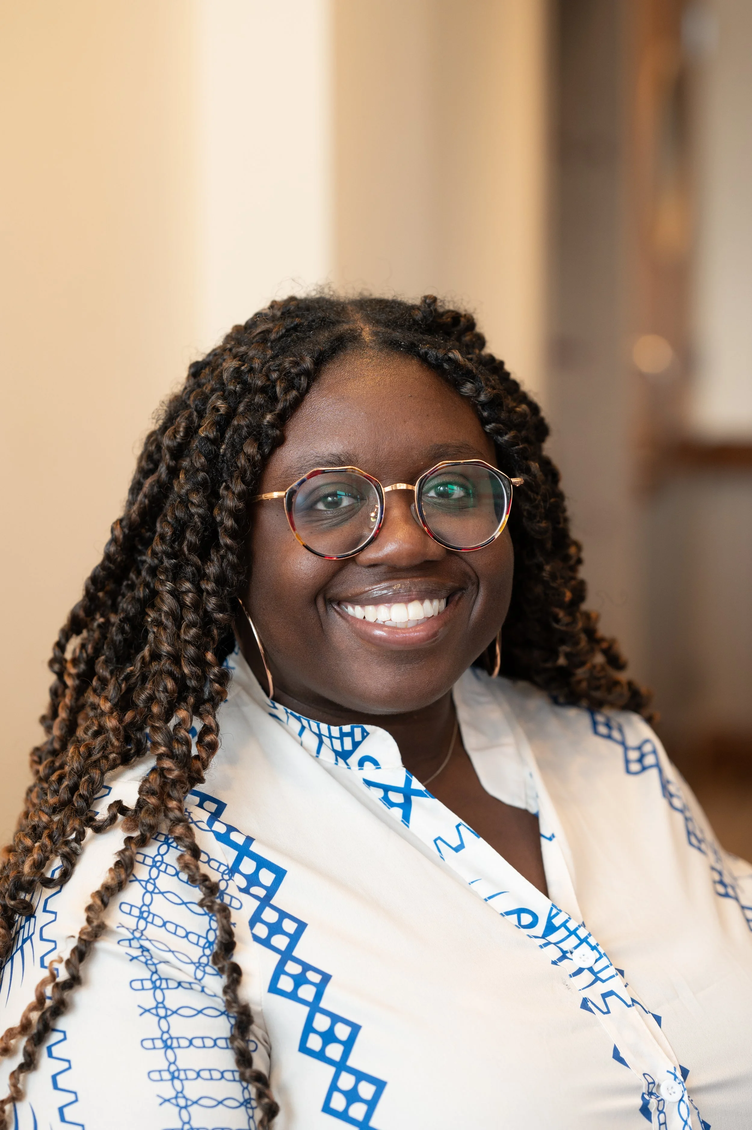 A woman with dark skin, long beaded braids, round glasses, and hoop earrings smiling at the camera.