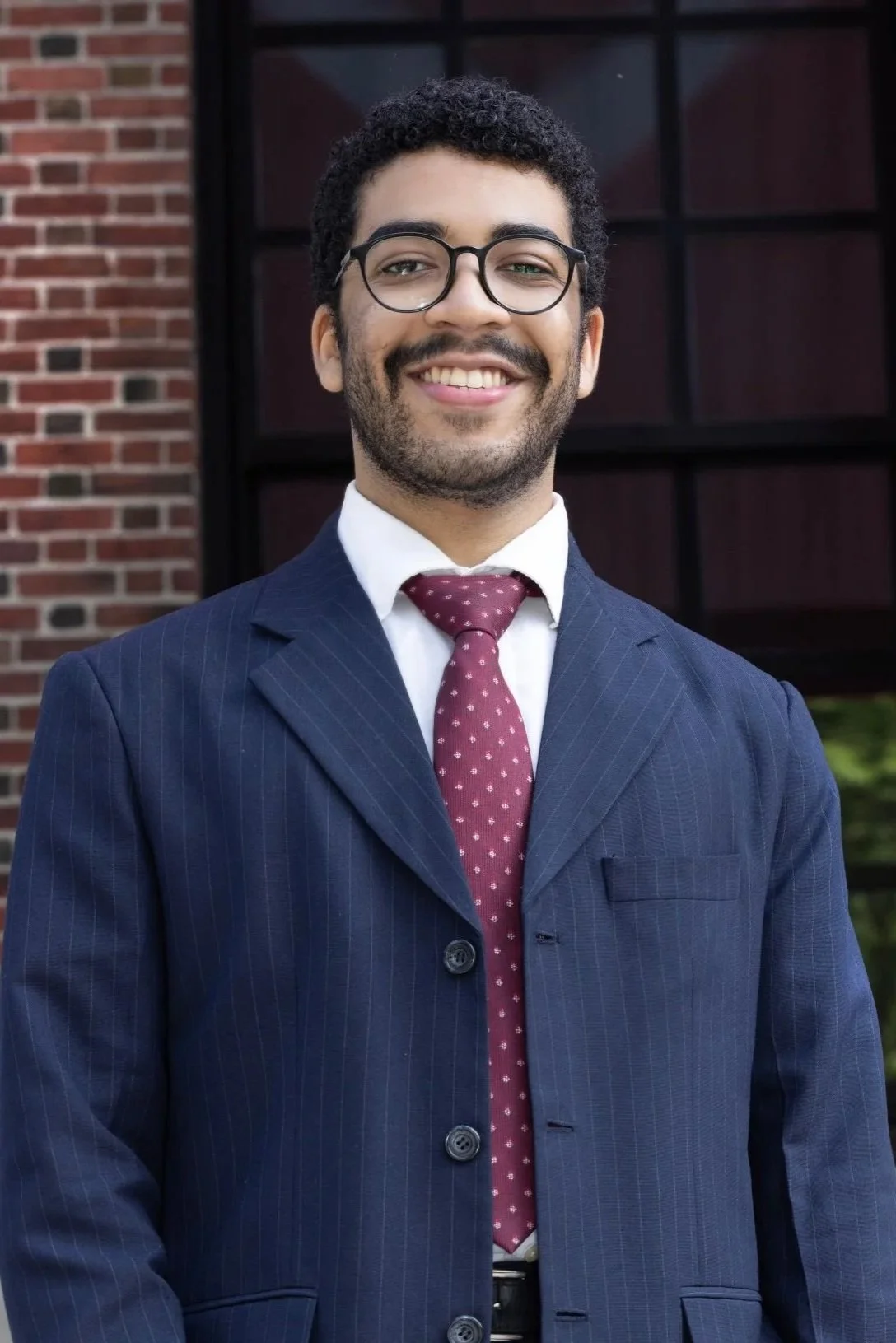 A young man with short, dark curly hair and light skin, smiling, wearing a dark blazer over a light blue button-up shirt, against a gray background.