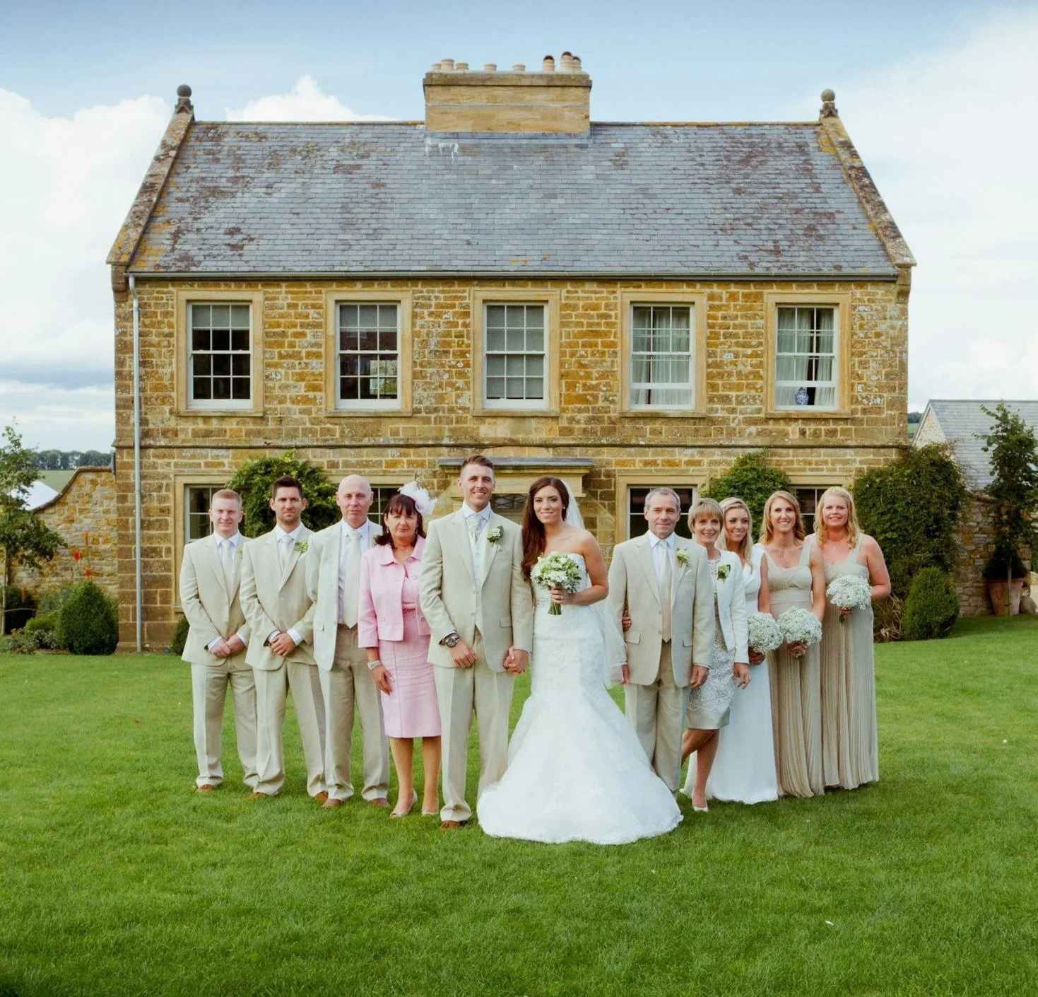 Group of wedding guests, including bride and groom, standing on grass in front of a stone house with four windows, during daytime.