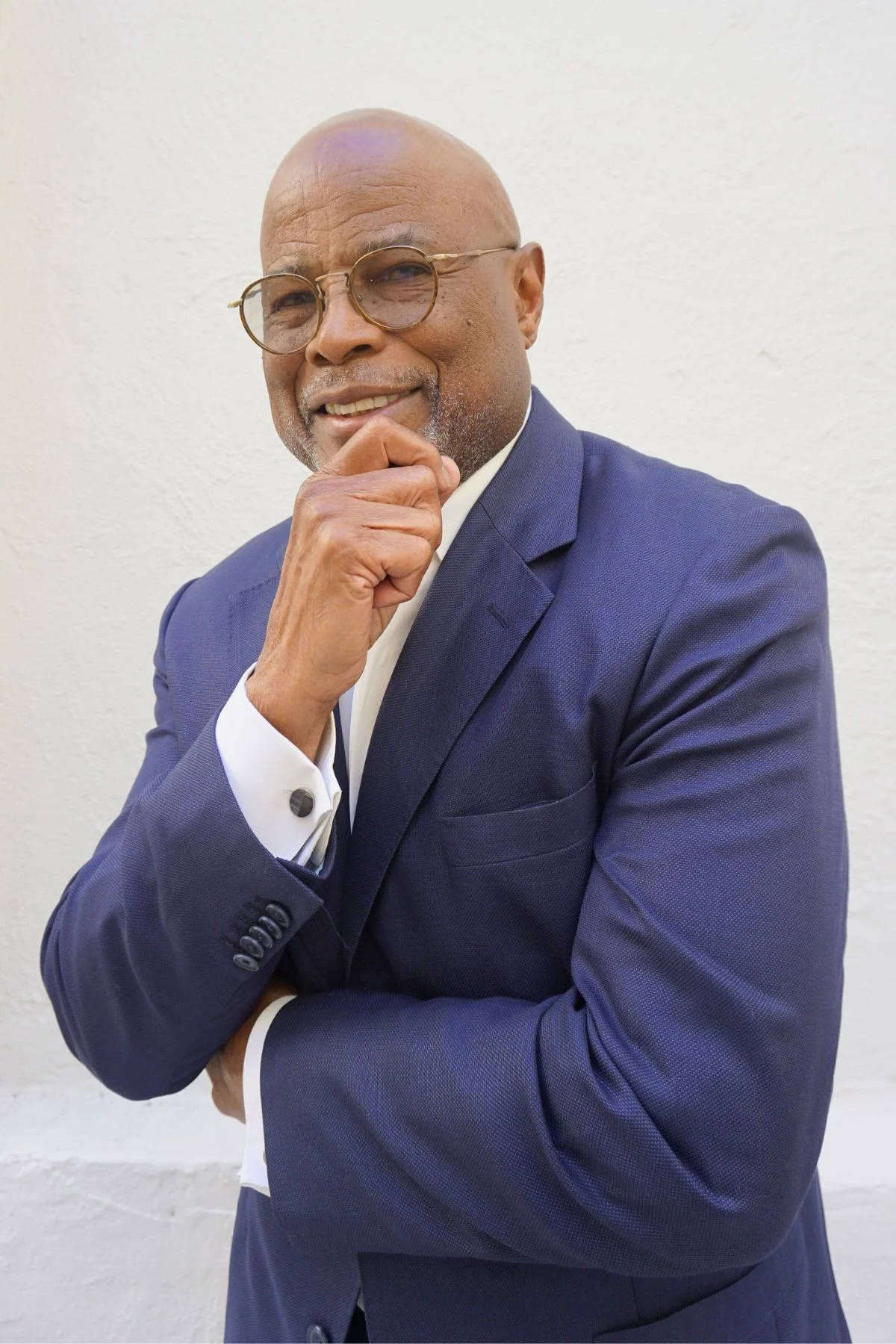 Portrait of a confident African American man in a blue suit and glasses, posing with hand on chin against a plain white wall.