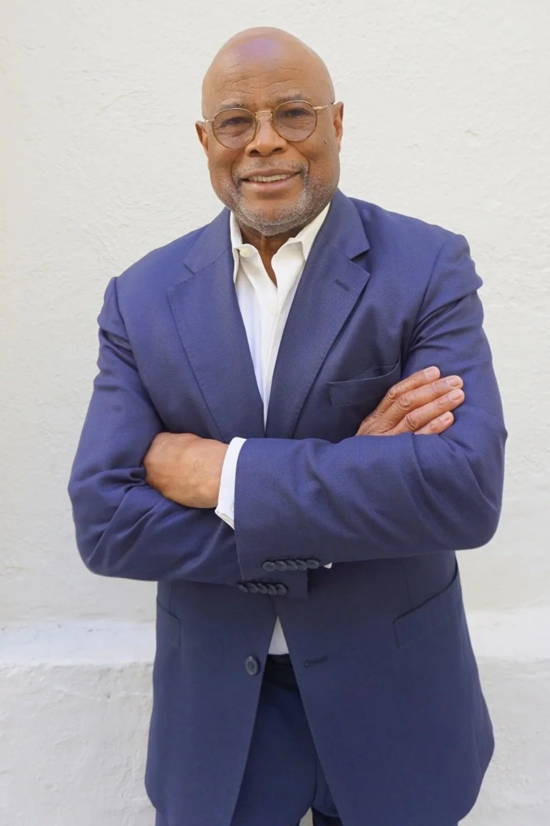 A middle-aged man with glasses, a beard, and a bald head is wearing a navy blue suit and white shirt, standing in front of a plain white wall with arms crossed and smiling.