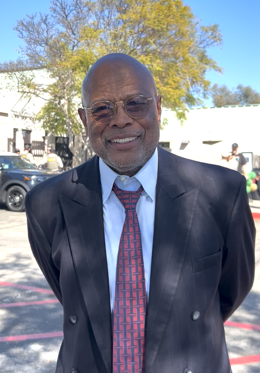 A smiling man in a dark pinstripe suit, white shirt, and patterned tie, wearing round glasses, outdoors with trees and cars in the background.