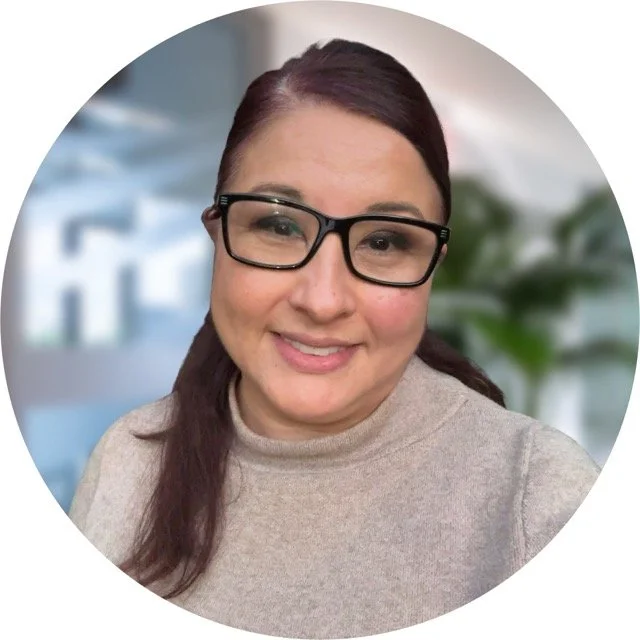 A woman with dark brown hair, wearing black glasses and a beige turtleneck sweater, smiling at the camera. The background is blurred with hints of blue and green.