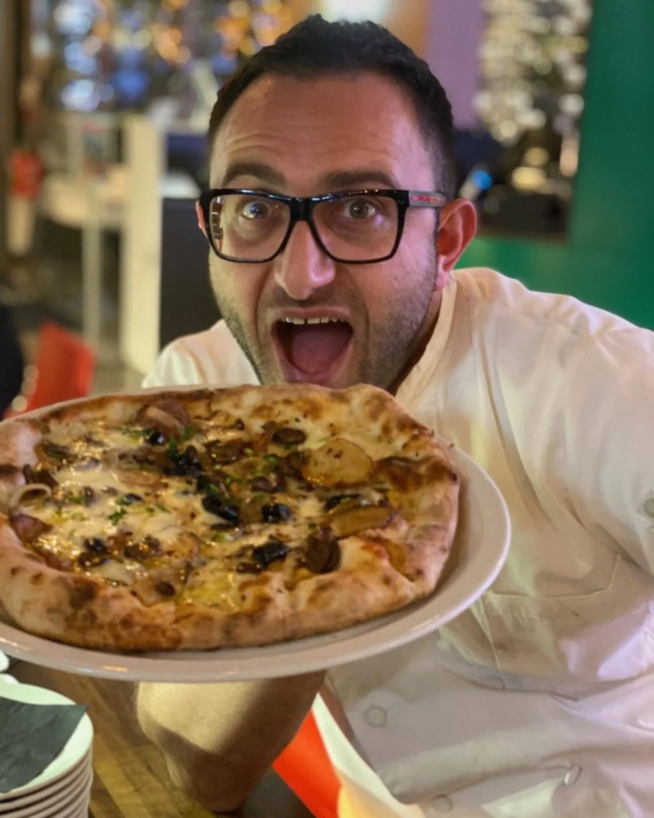 Man with glasses excitedly holding a plate with a pizza topped with vegetables and mushrooms in a restaurant setting.