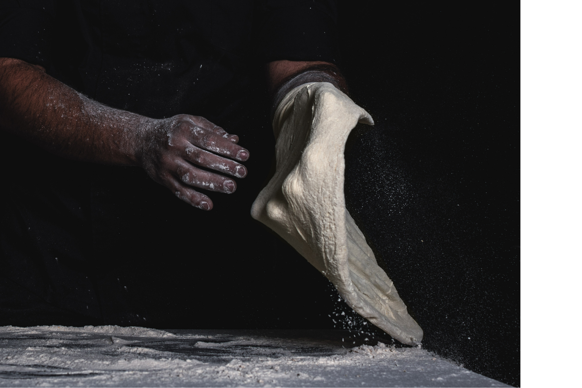 A baker's hands tossing pizza dough in the air, with flour in the air and on the surface, against a dark background.