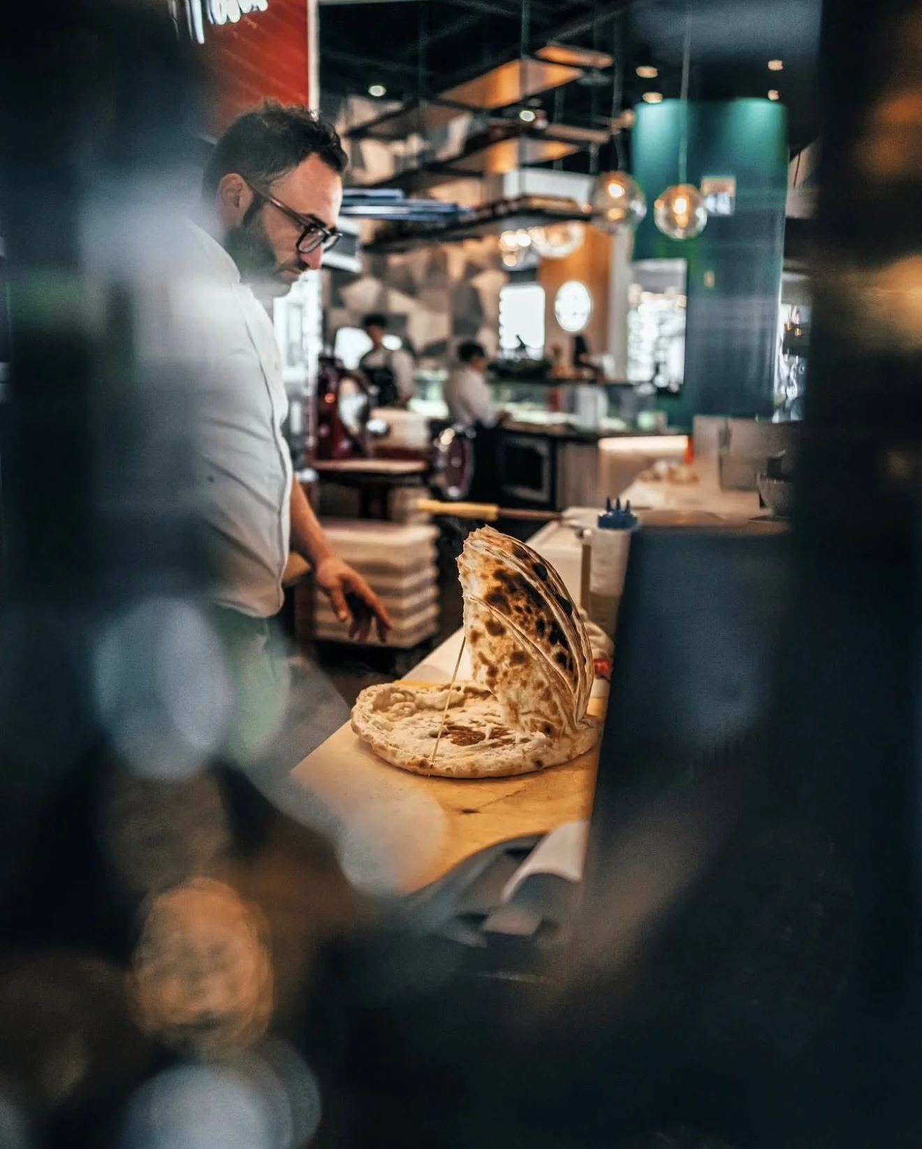 A chef preparing flatbread or pizza in a modern restaurant kitchen with warm lighting and blurred background.