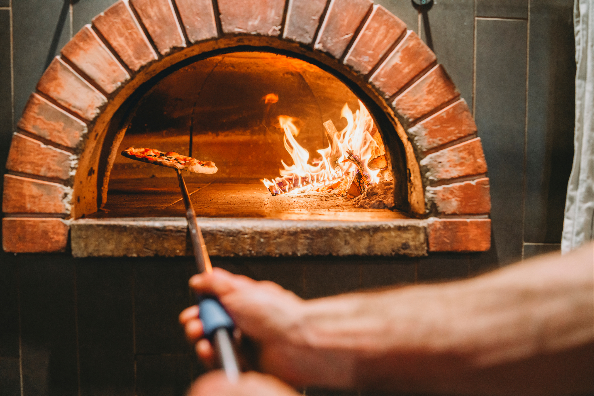Person using a pizza peel to place or remove a pizza from a wood-fired brick oven with flames inside.