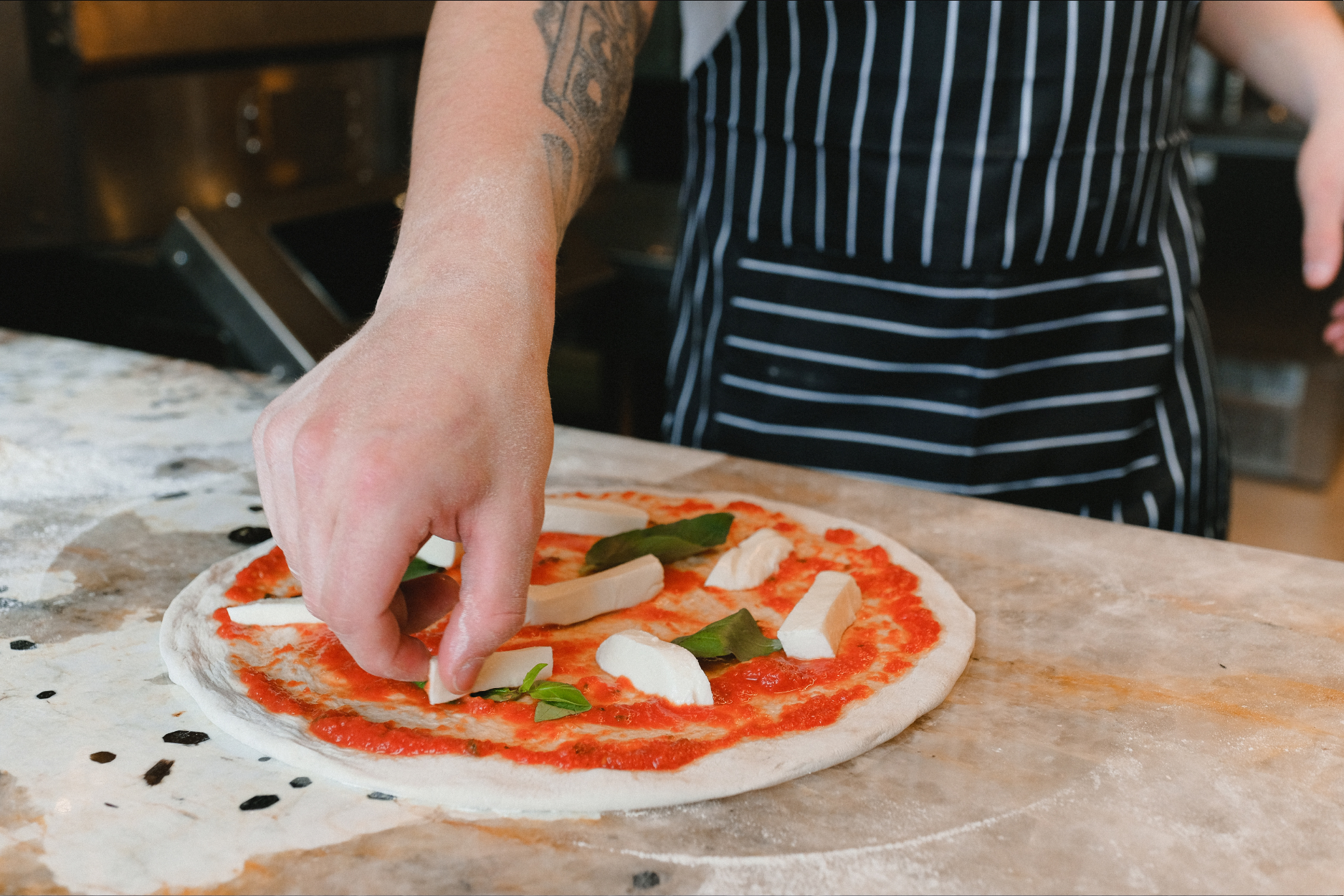 Person placing mozzarella cheese and basil leaves on a pizza dough with tomato sauce.