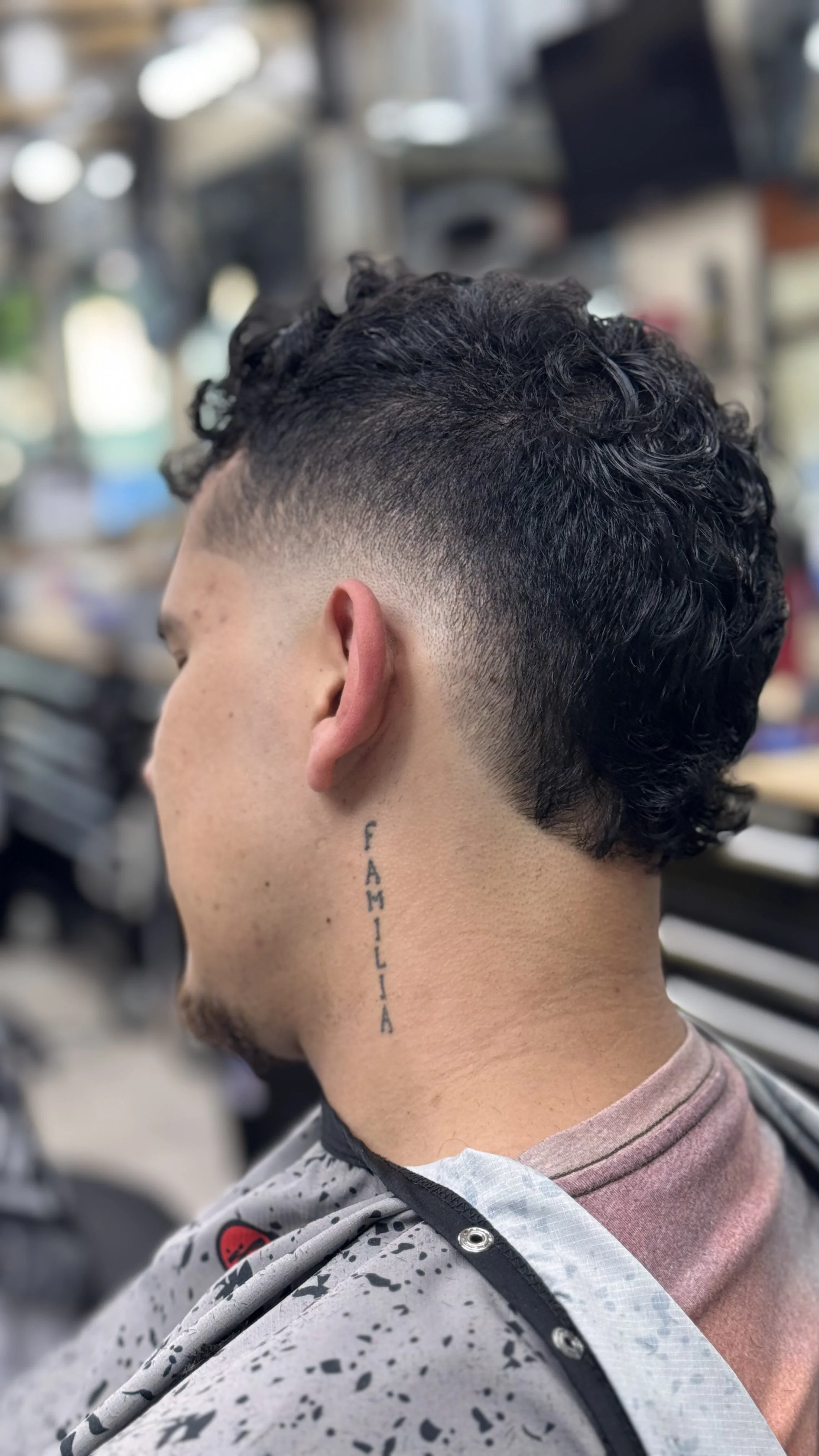 Side view of a young man with a fresh, curly undercut hairstyle and the word 'FAMILIA' tattooed vertically on his neck.