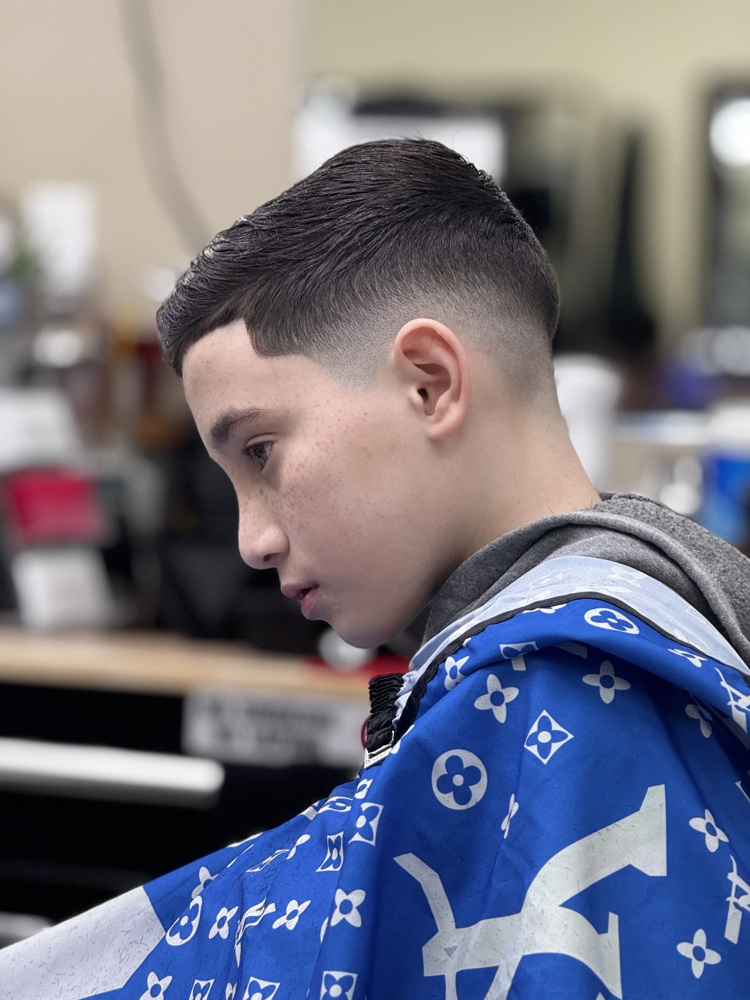 Young man with a short, clean haircut, sitting in a barbershop. He's wearing a blue Louis Vuitton barber cape and looking down, possibly reading or in thought. The background is blurred with typical barbershop equipment.