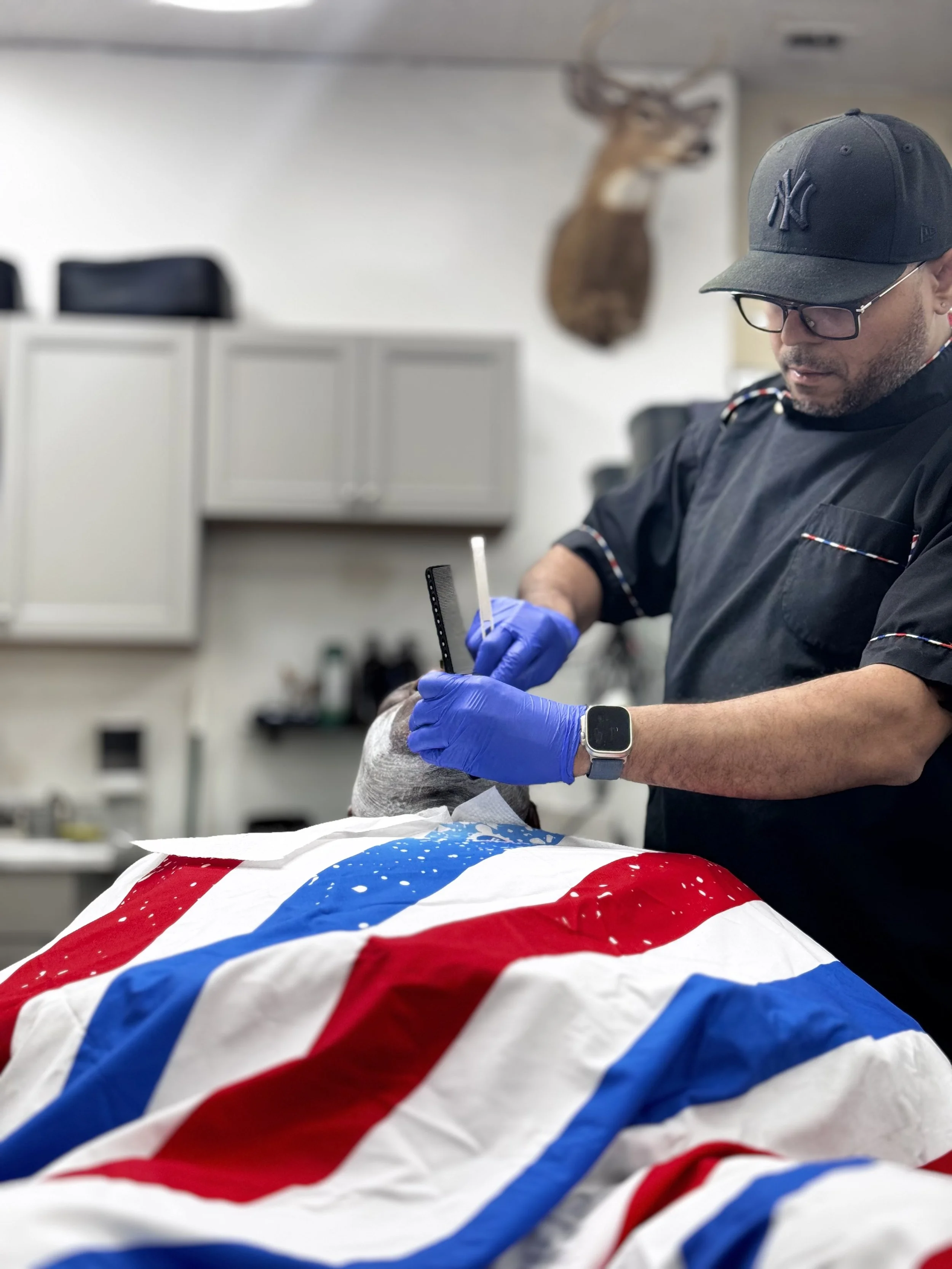 A man wearing glasses, a baseball cap, and a black uniform is tattooing a person's face with a tattoo needle. The person receiving the tattoo is lying down covered with a fabric featuring a red, white, and blue pattern. A mounted deer head is visible on the wall in the background.