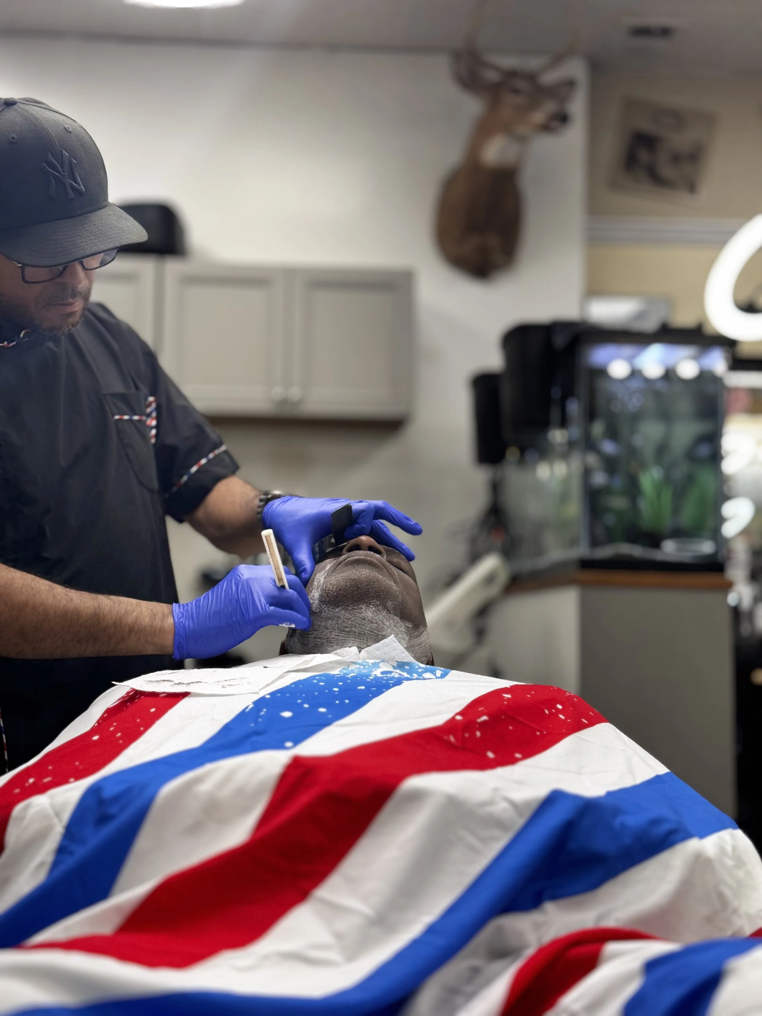A barber, wearing glasses, a black NY Yankees cap, and blue gloves, is applying shave cream to a man lying back with a barber cape featuring red, white, and blue stripes, in a barber shop with a deer head mounted on the wall in the background.