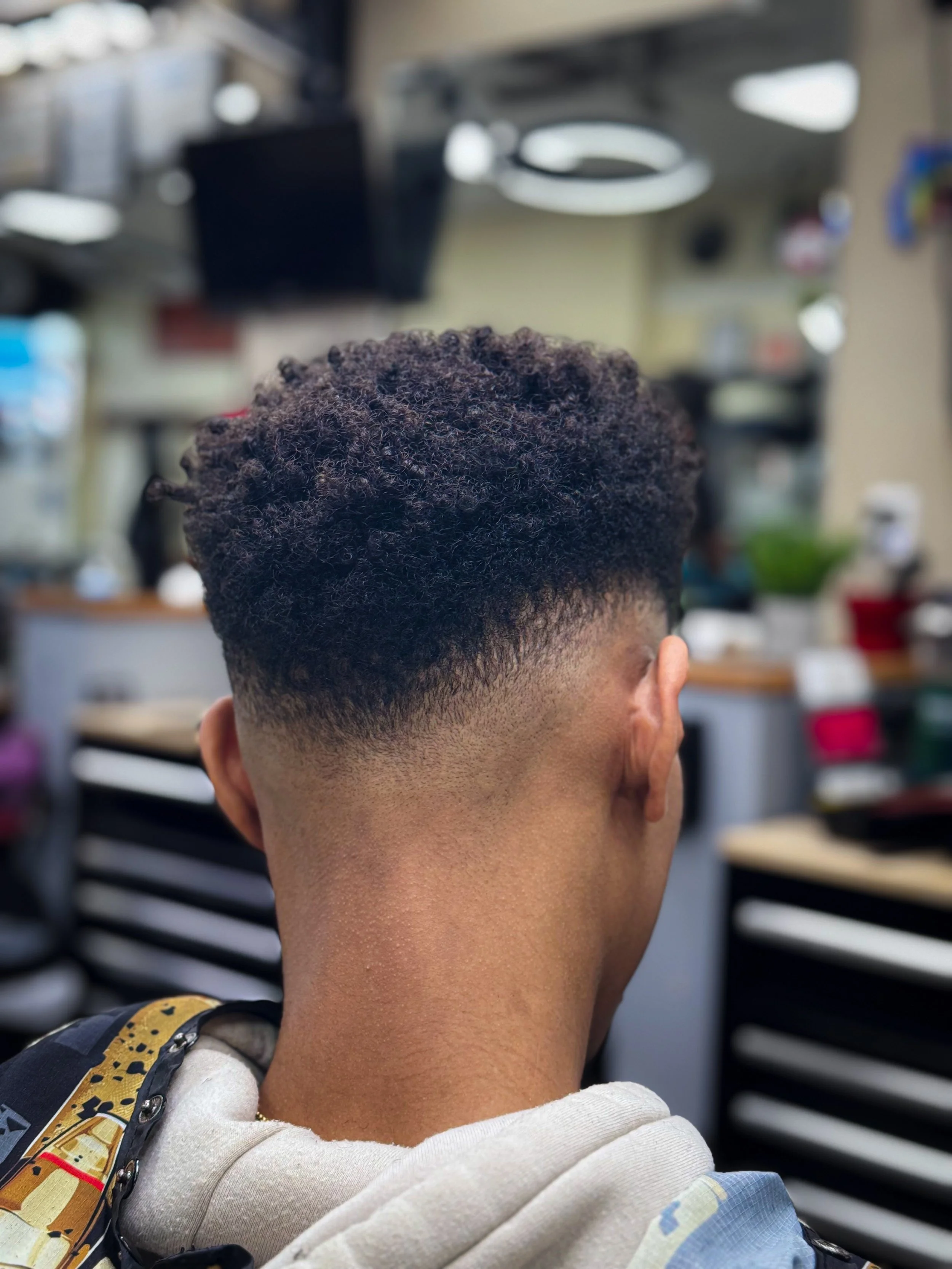 Back of a person's head showing a fade haircut with curly hair on top, in a barbershop setting.