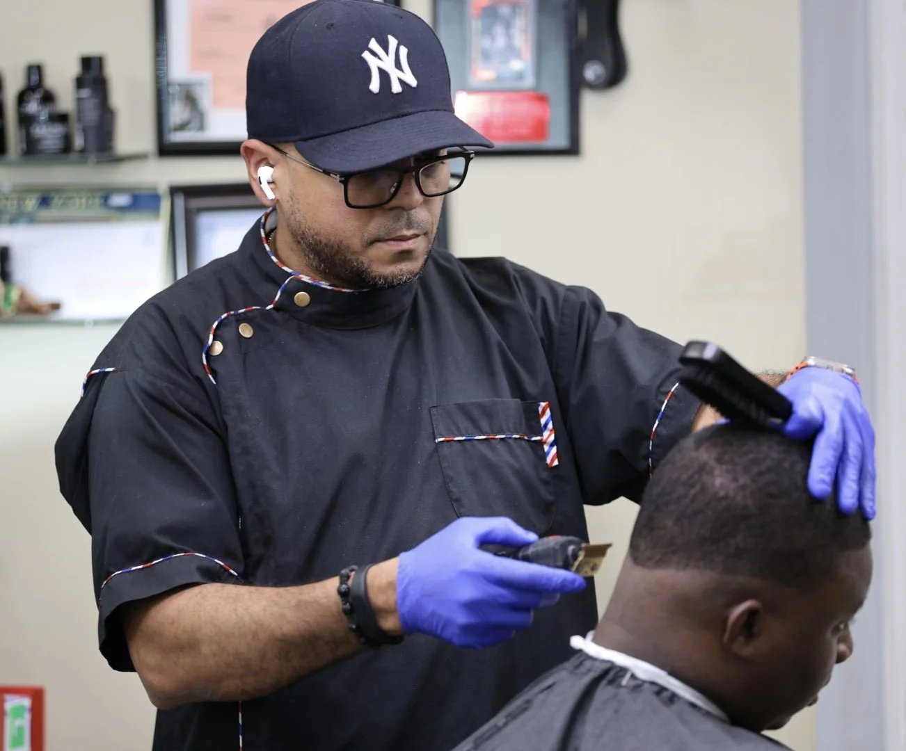 Barber wearing a Navy Yankees cap, glasses, and blue gloves gives a haircut to a client in a barbershop.