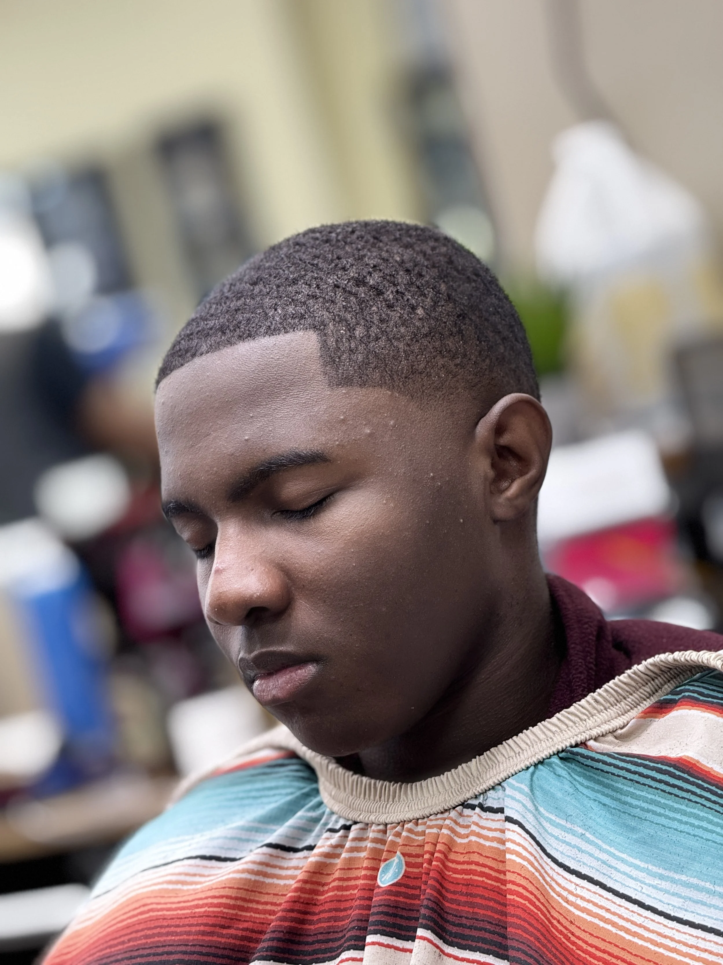 A young African American man with a fresh fade haircut and closed eyes, sitting in a barbershop wearing a multicolored striped cape.