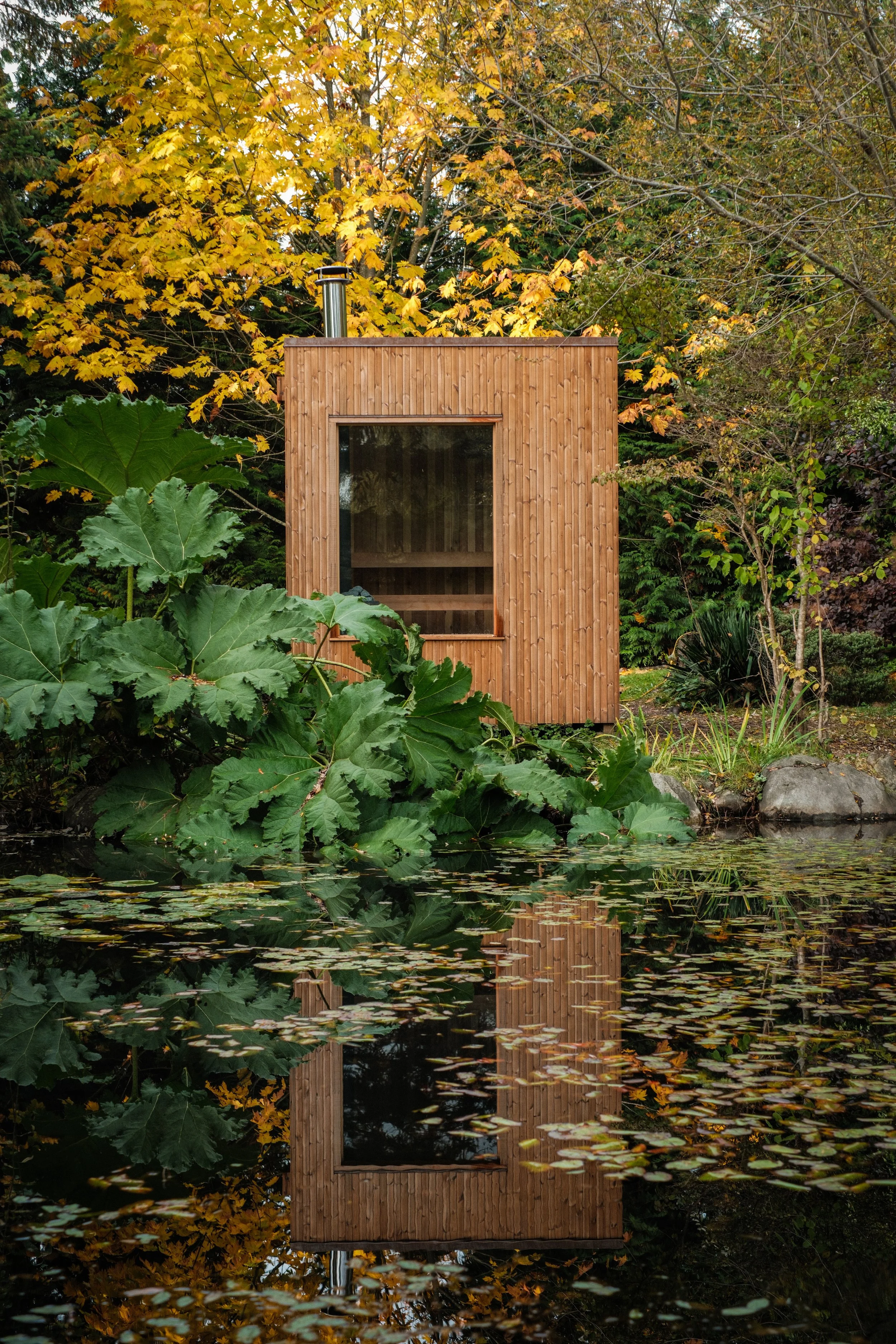 A large, modern wooden sauna with large windows is situated by a pond surrounded by lush green and autumn-colored foliage, with its reflection visible in the water.