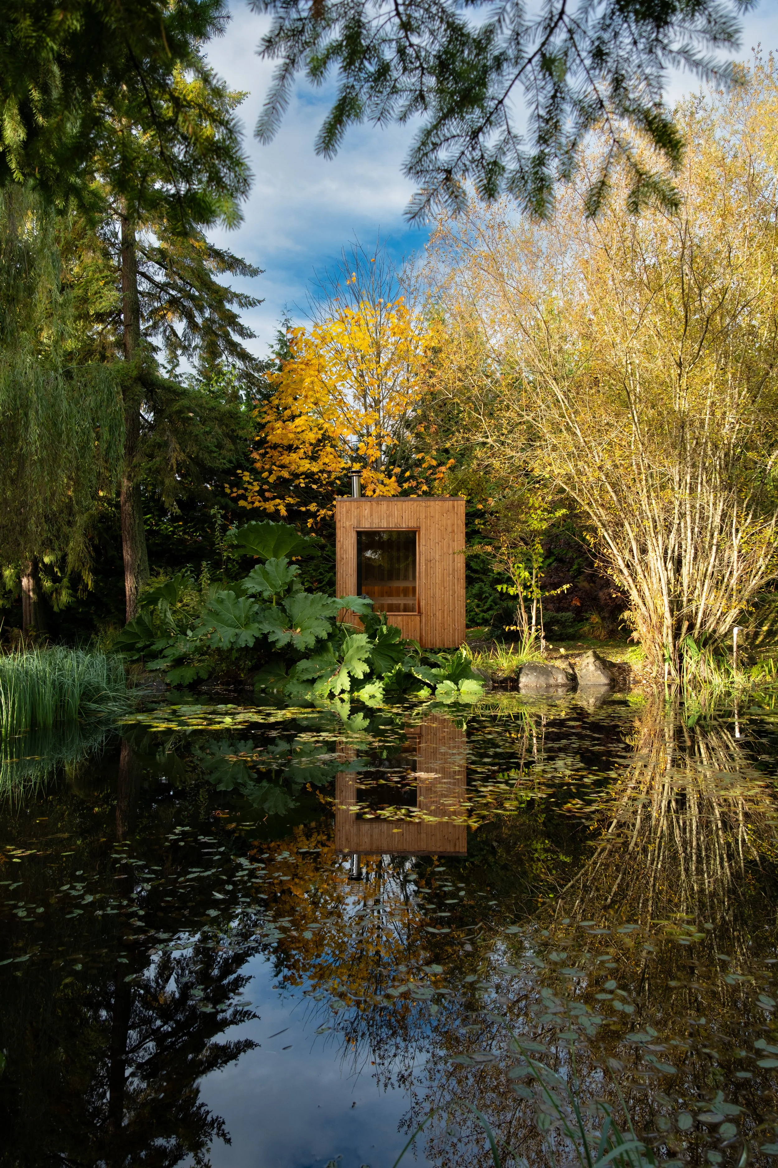 A large wooden sauna on the edge of a pond surrounded by trees with yellow and green leaves, reflecting in the water under a partly cloudy sky.