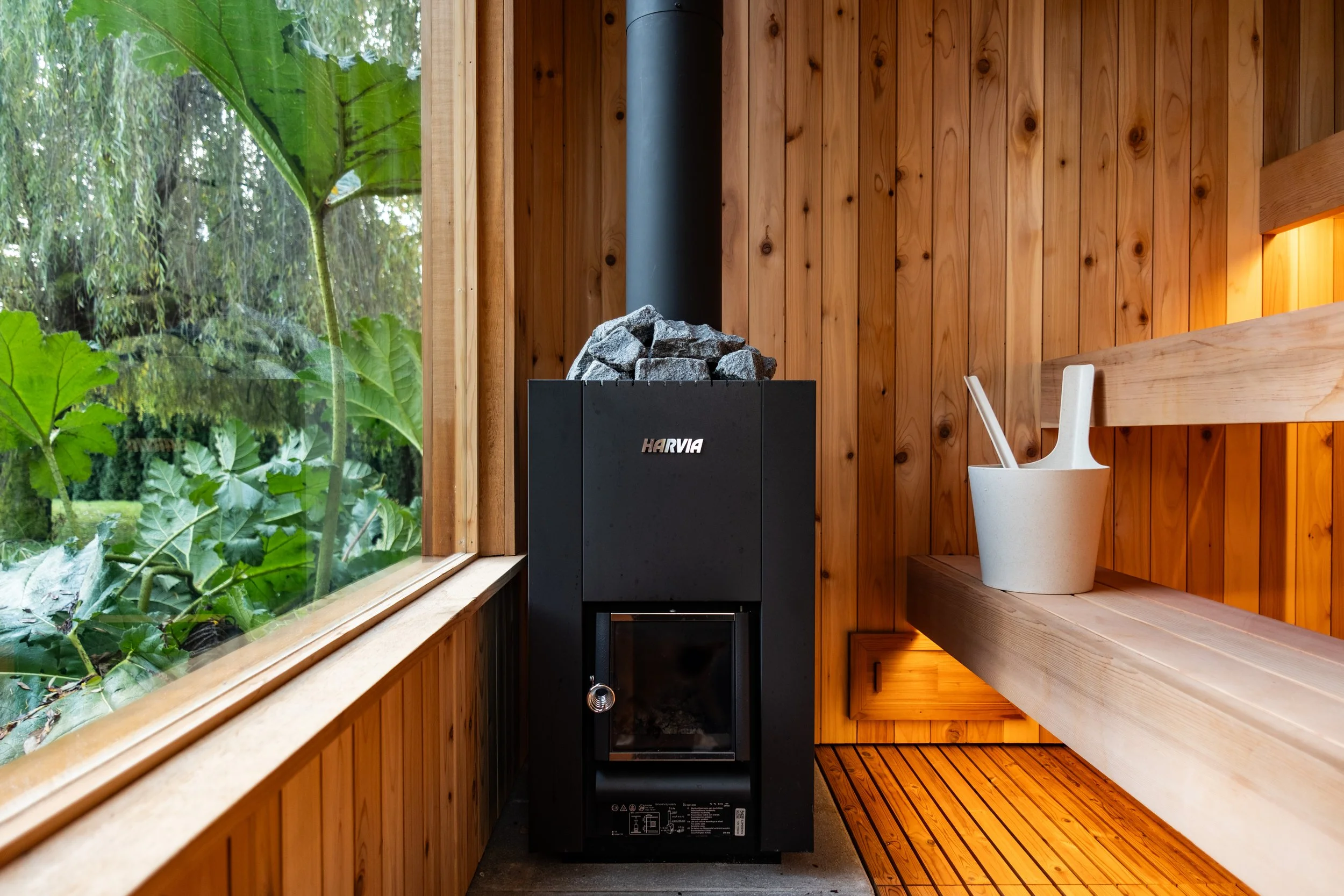 Inside a sauna with wooden walls, a heater with rocks, a large window showing green trees and leaves, and a wooden bench with a white bucket and sauna accessories.