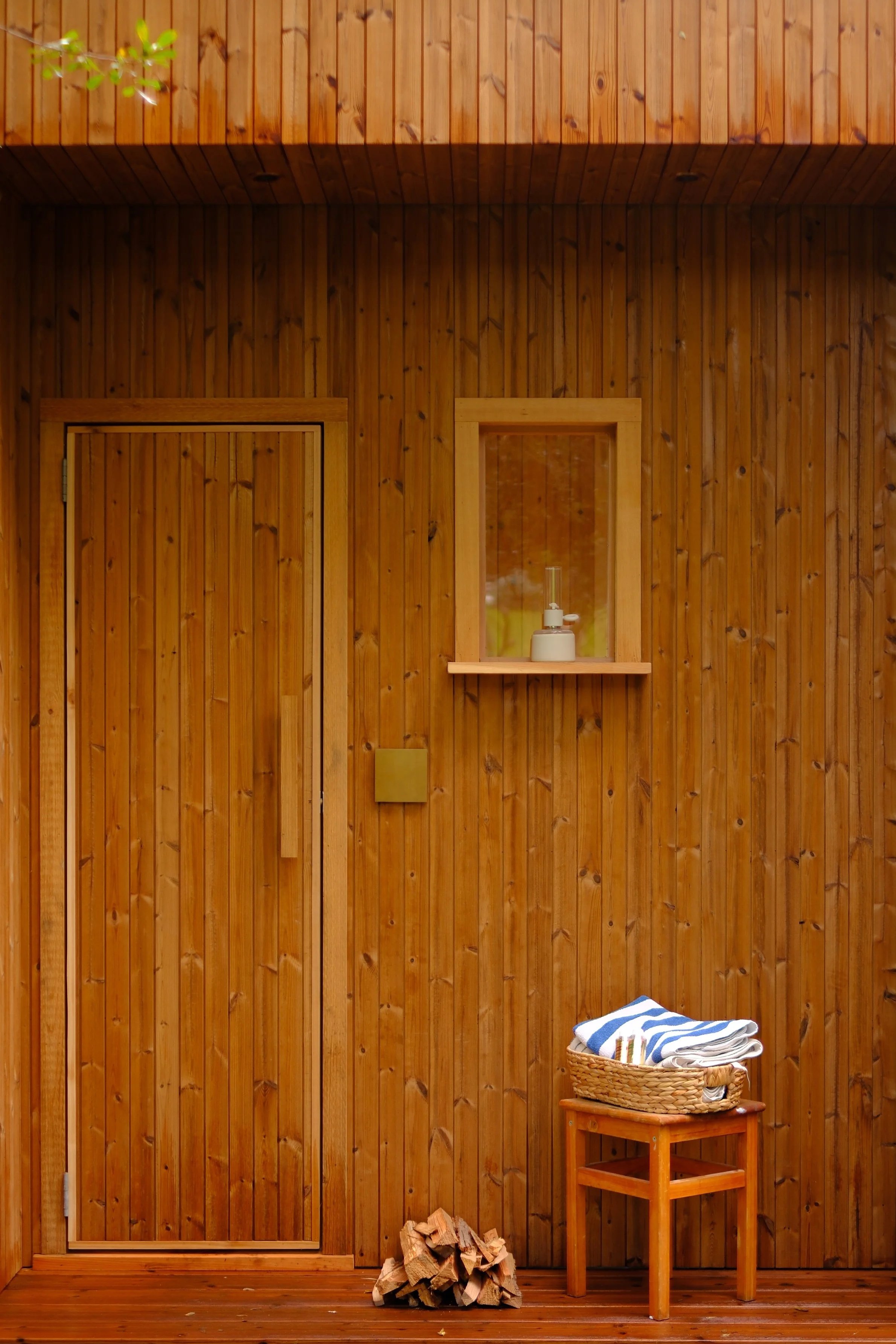 Front of a wooden sauna with a closed door, a small window with a sauna lamp, a basket of towels on a small wooden table, and firewood on the floor.