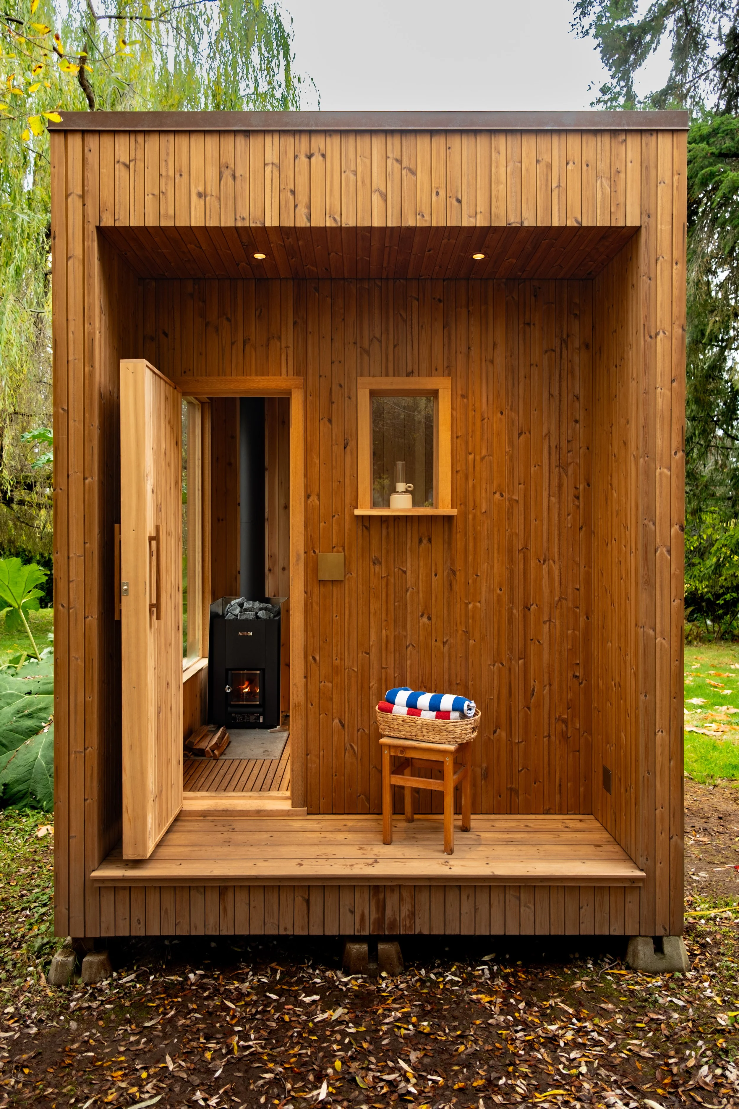 Wooden outdoor sauna with open door, small window with shelf, striped towel in basket, and wood stove inside, set in a natural outdoor environment.