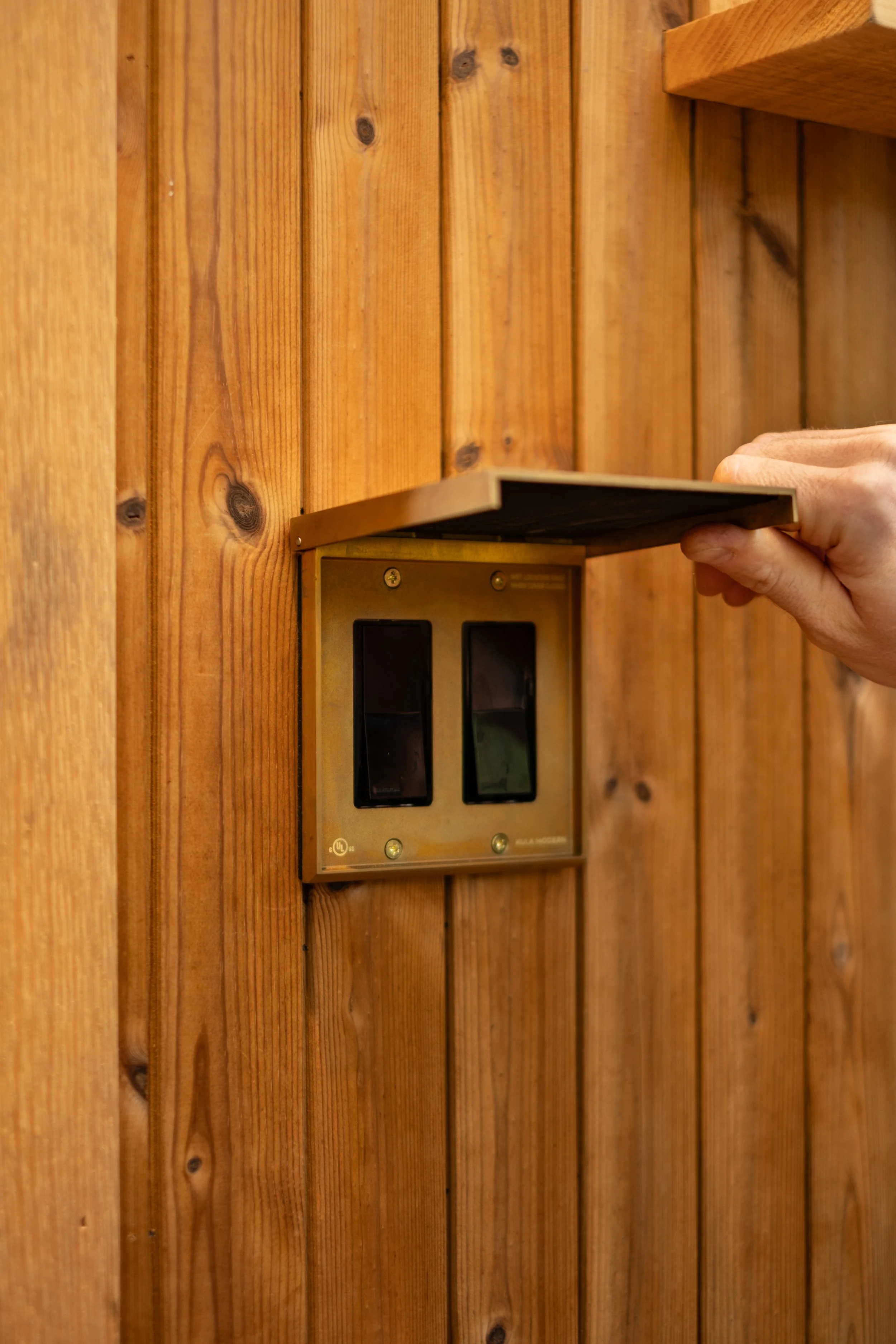 Person opening a brass light switch cover on a wooden wall.