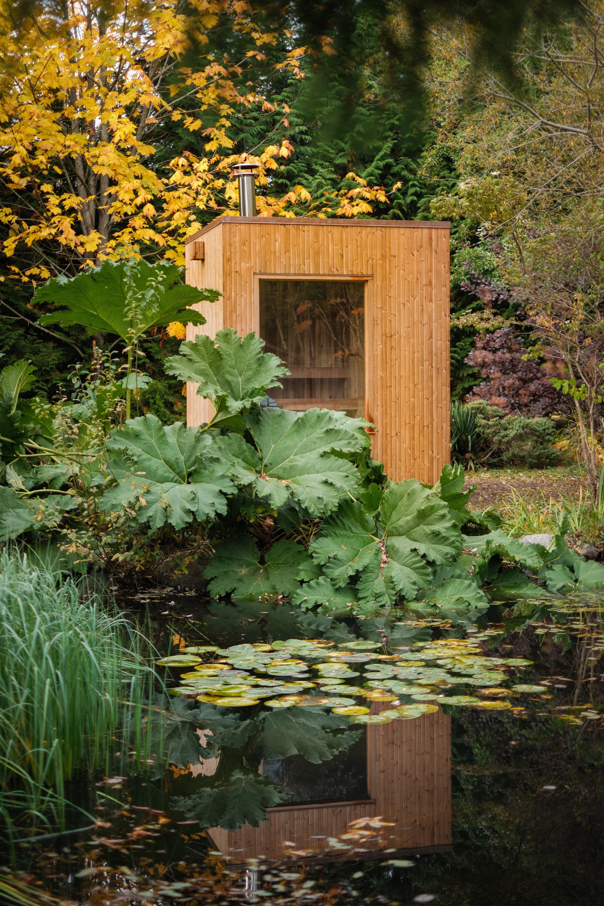 A large wooden sauna beside a pond with lily pads, surrounded by trees with autumn foliage.