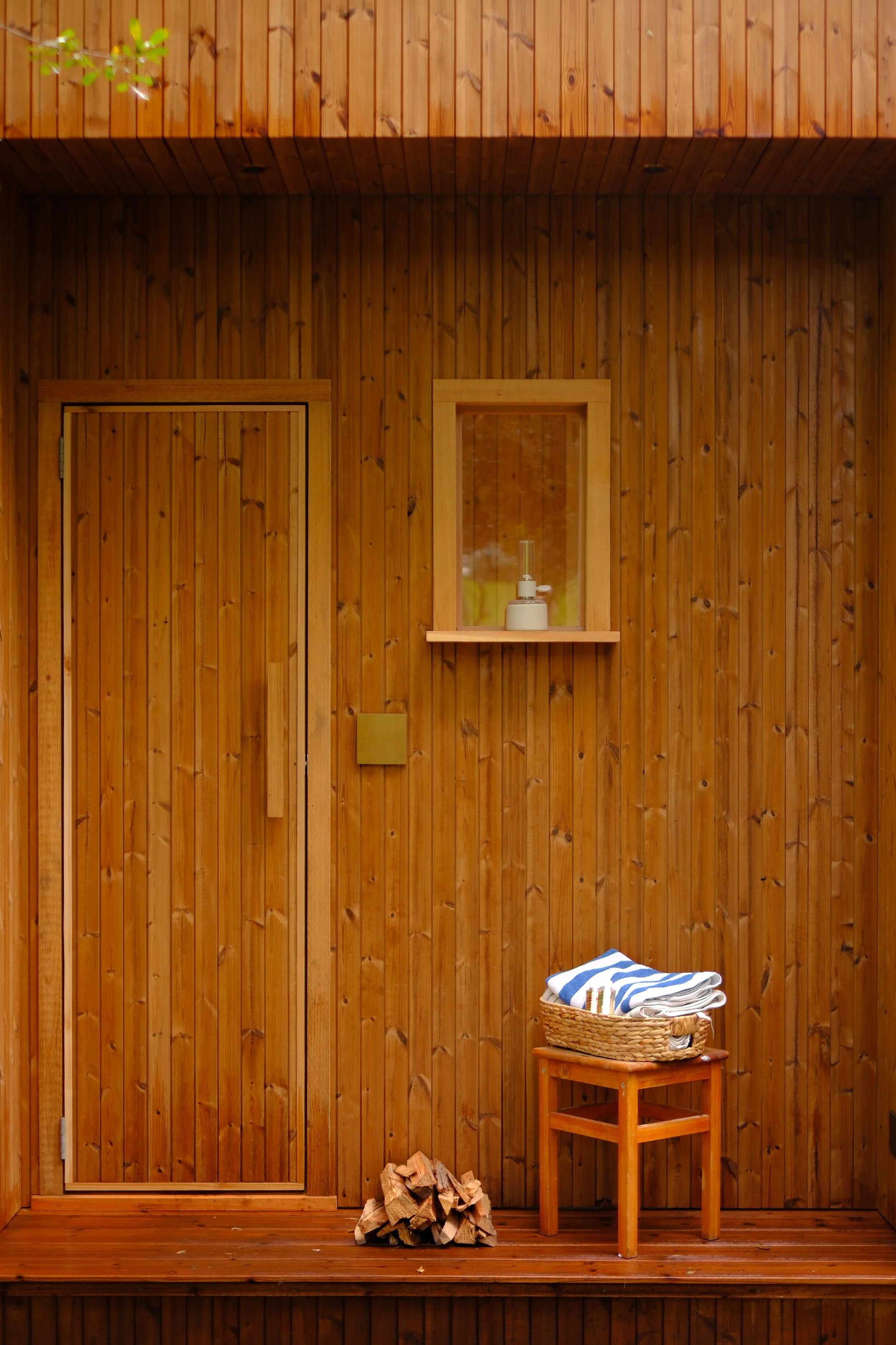 Indoor wooden sauna with stacked firewood on the floor, a small wooden table with folded towels, a basket, and a window with a soap dispenser on the shelf.