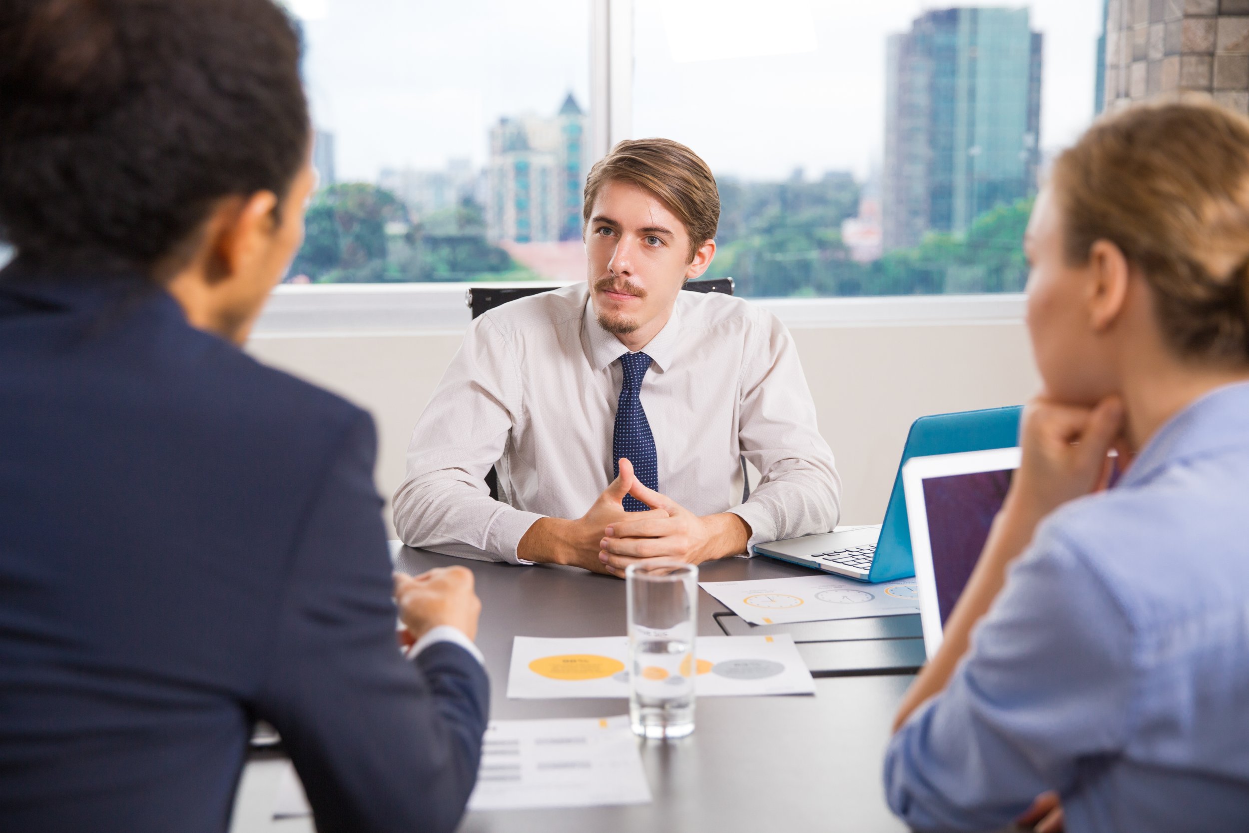 Three people in business attire having a meeting in a modern office with a city view. The man in the center is speaking, seated at a table with documents, a laptop, and a glass of water.