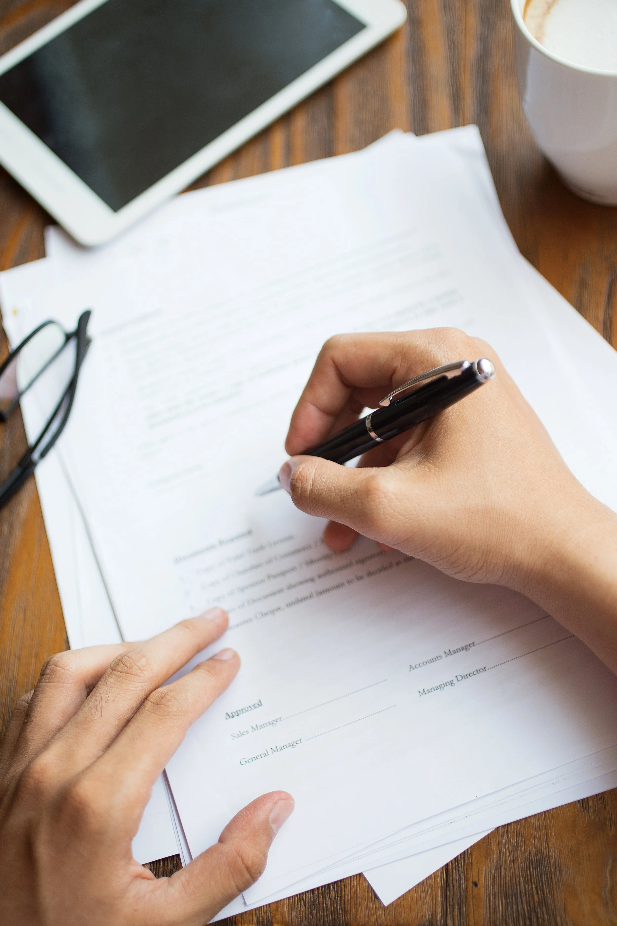 Person signing a document at a wooden table with glasses, a tablet, and a cup of coffee nearby.