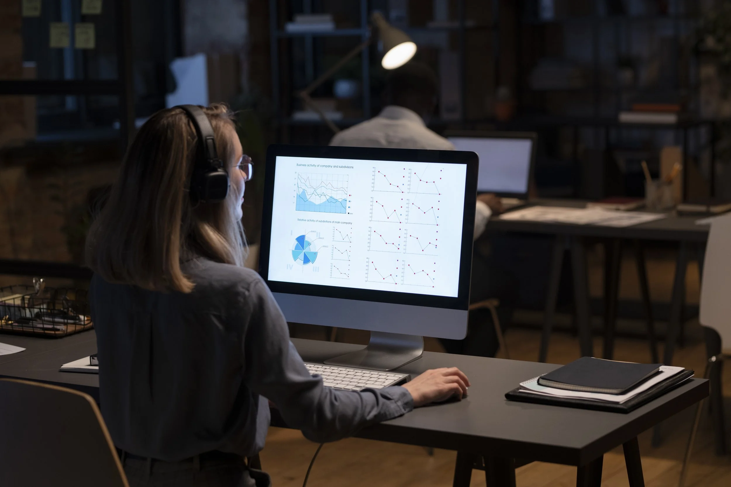 Woman wearing headphones working on a computer displaying charts and graphs in a dimly lit office.