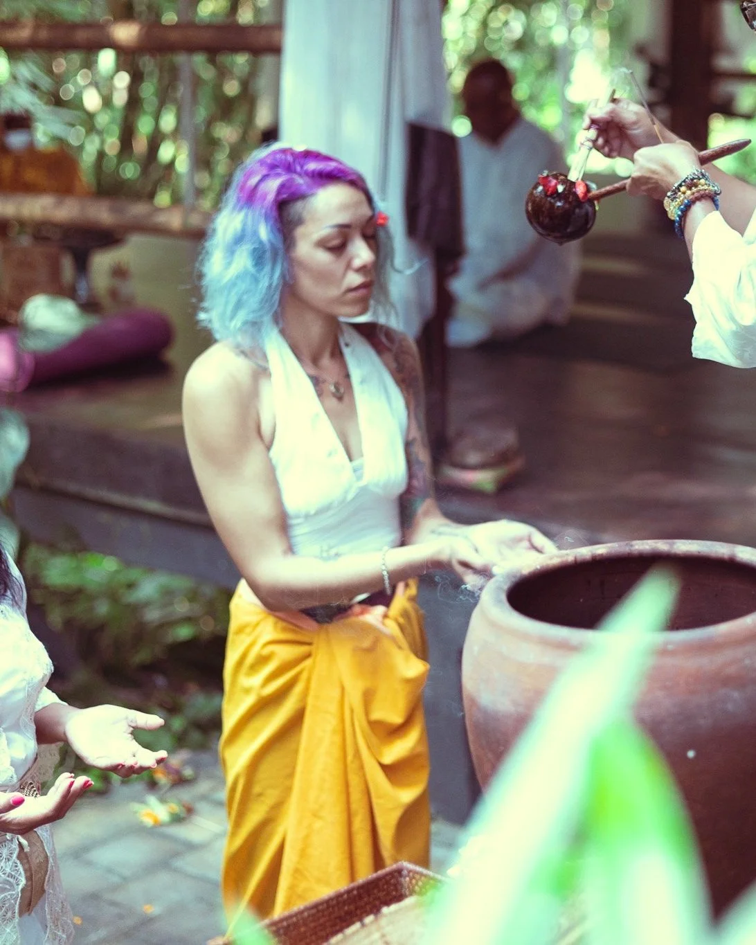 A woman with colorful purple and blue hair and tattoos is participating in a spiritual or ritual activity, with her hands cupped above a large clay pot, while another person holds a small vessel above her. The setting appears to be outdoors in a lush, green environment.