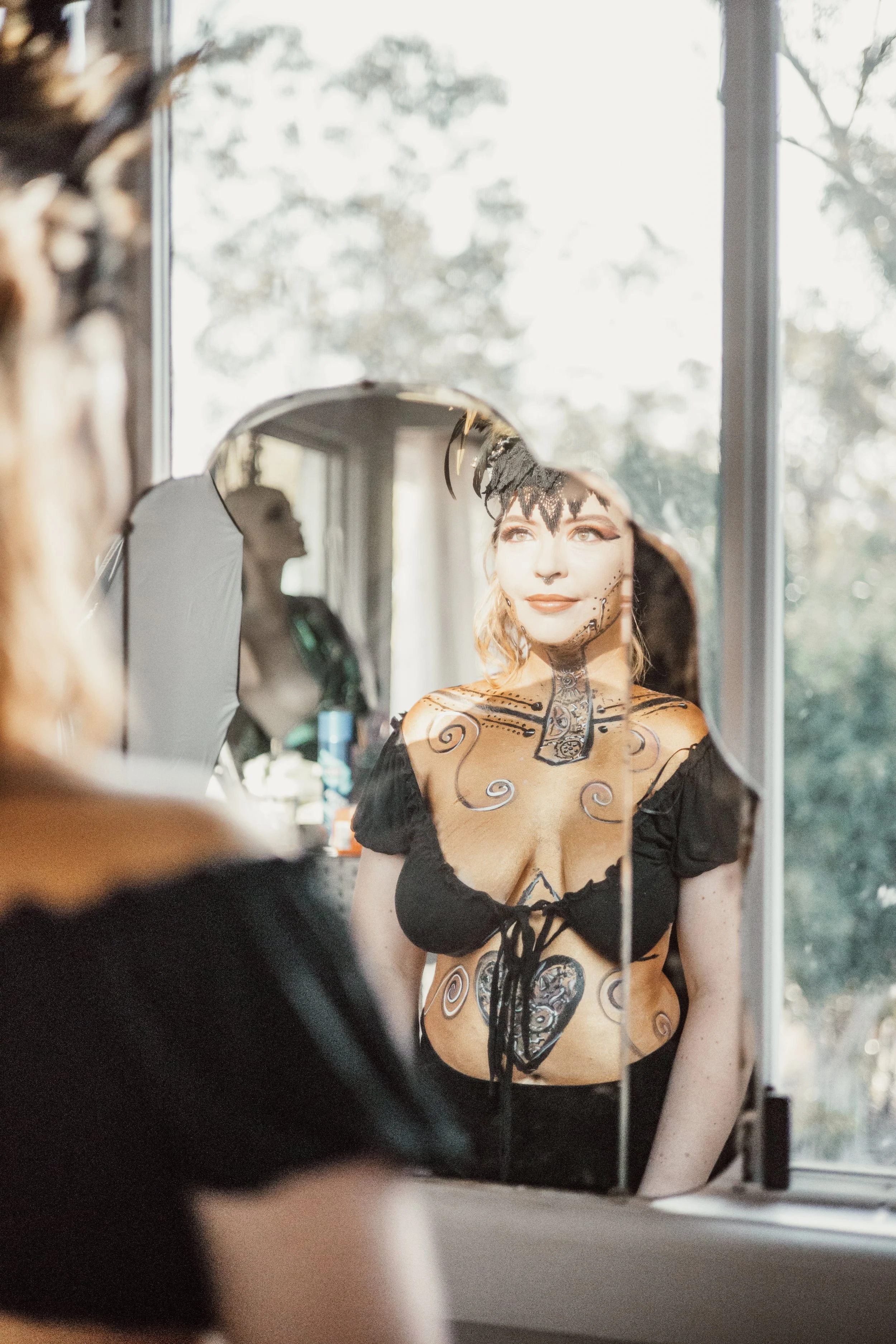 Woman with face paint looking at herself in a mirror, dressed in a black top with laced neckline, her face painted to resemble a skeleton or Dia de los Muertos theme, with feathered headpiece, in a well-lit room with large windows overlooking trees.