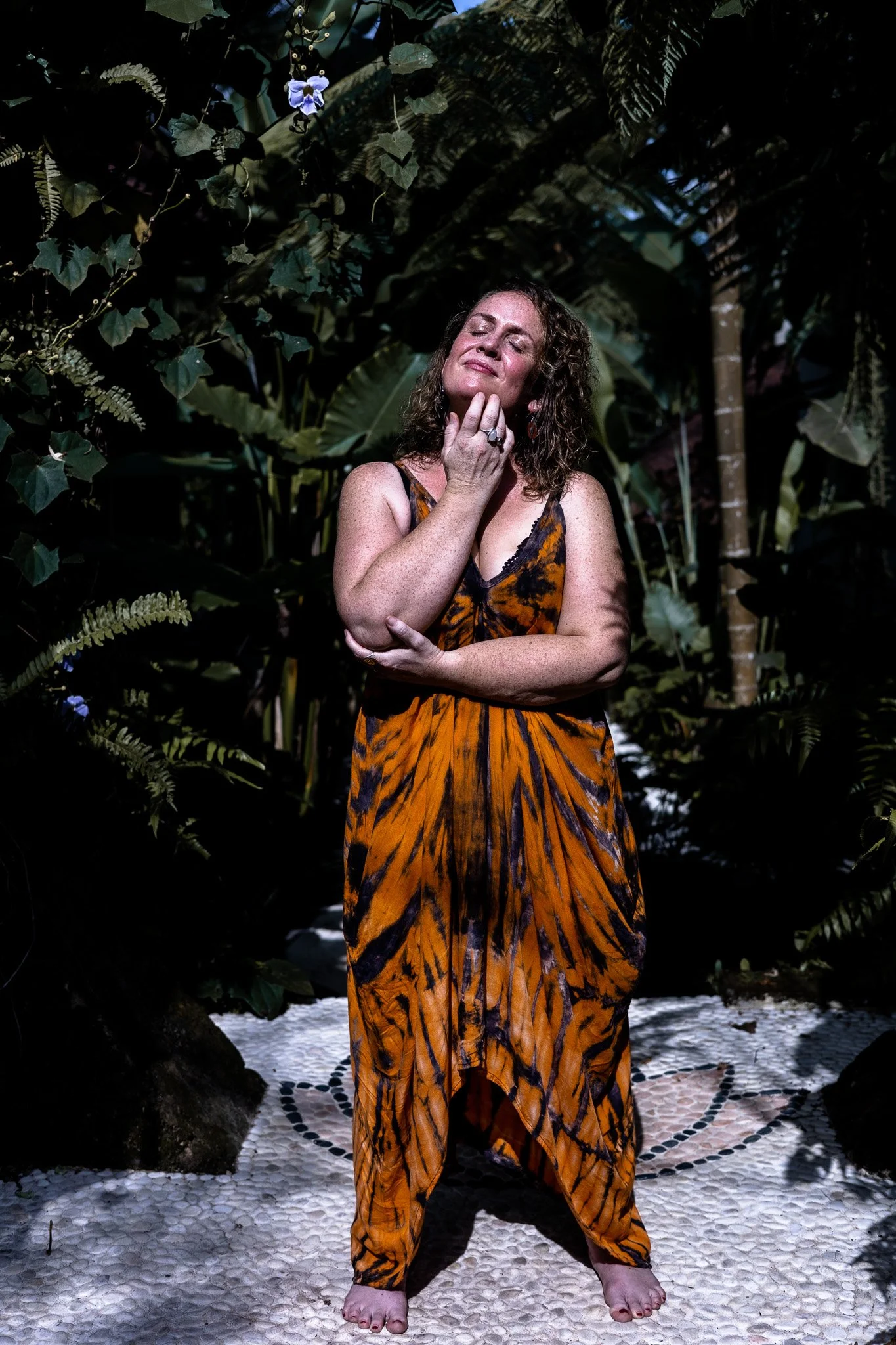 A woman in a tiger-stripe dress standing on a mosaic pathway surrounded by dense green tropical foliage, with her eyes closed and one hand touching her face in a thoughtful pose.