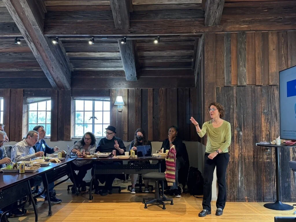A woman giving a presentation in a wooden conference room with several people seated at a long table, some eating and drinking, and a large screen to her right.