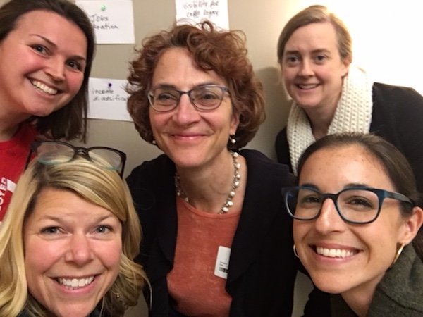 Five women smiling for a group selfie indoors, with papers on the wall behind them.